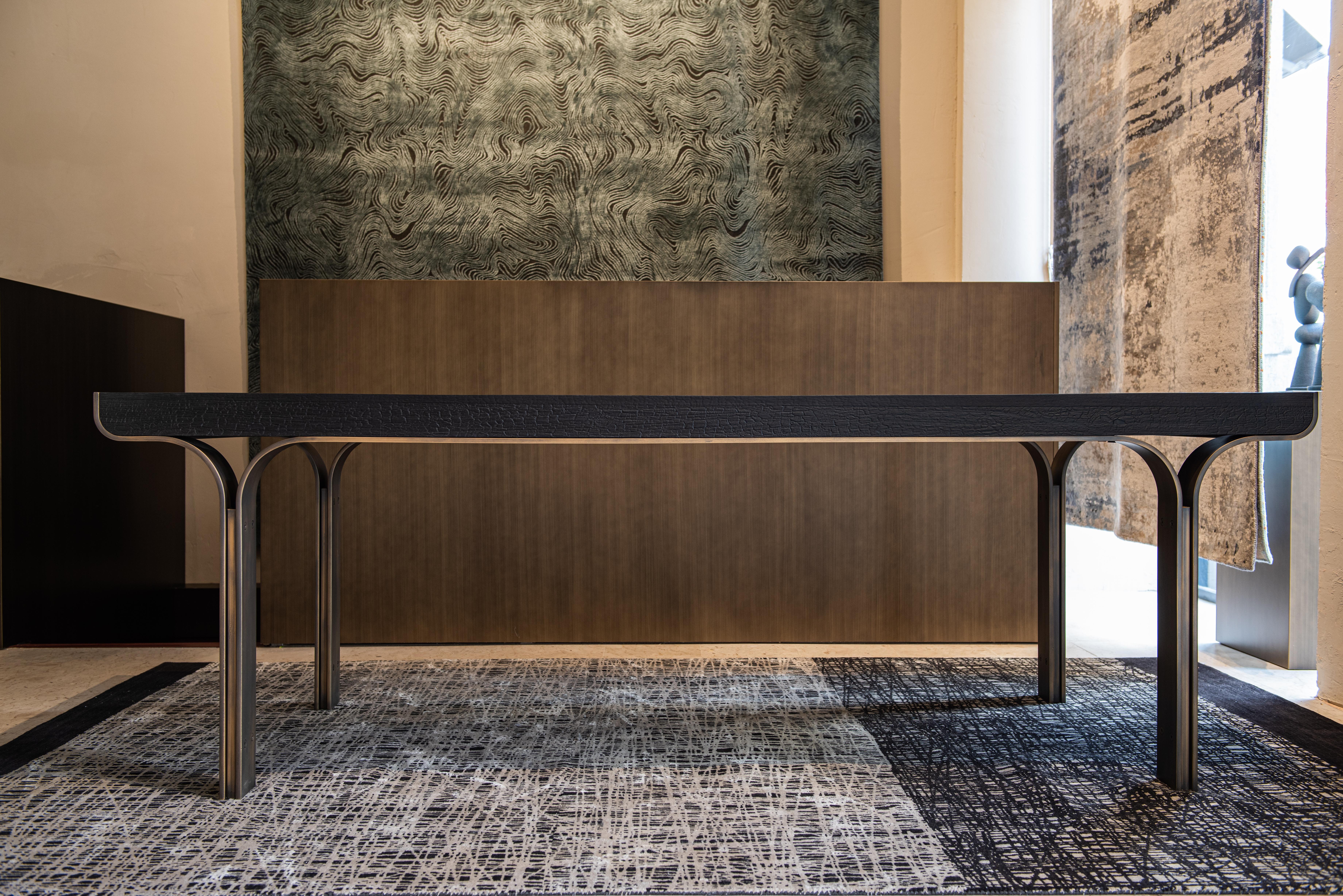 Osvaldo dining table by Delvis Unlimited.
Dimensions: D 90 x W 240 x H 76 cm.
Material: black burnt wood, burnished iron brassed.

The Osvaldo table, designed by Fabio Fantolino, is like a dance of shapes and materials. The steel structure,