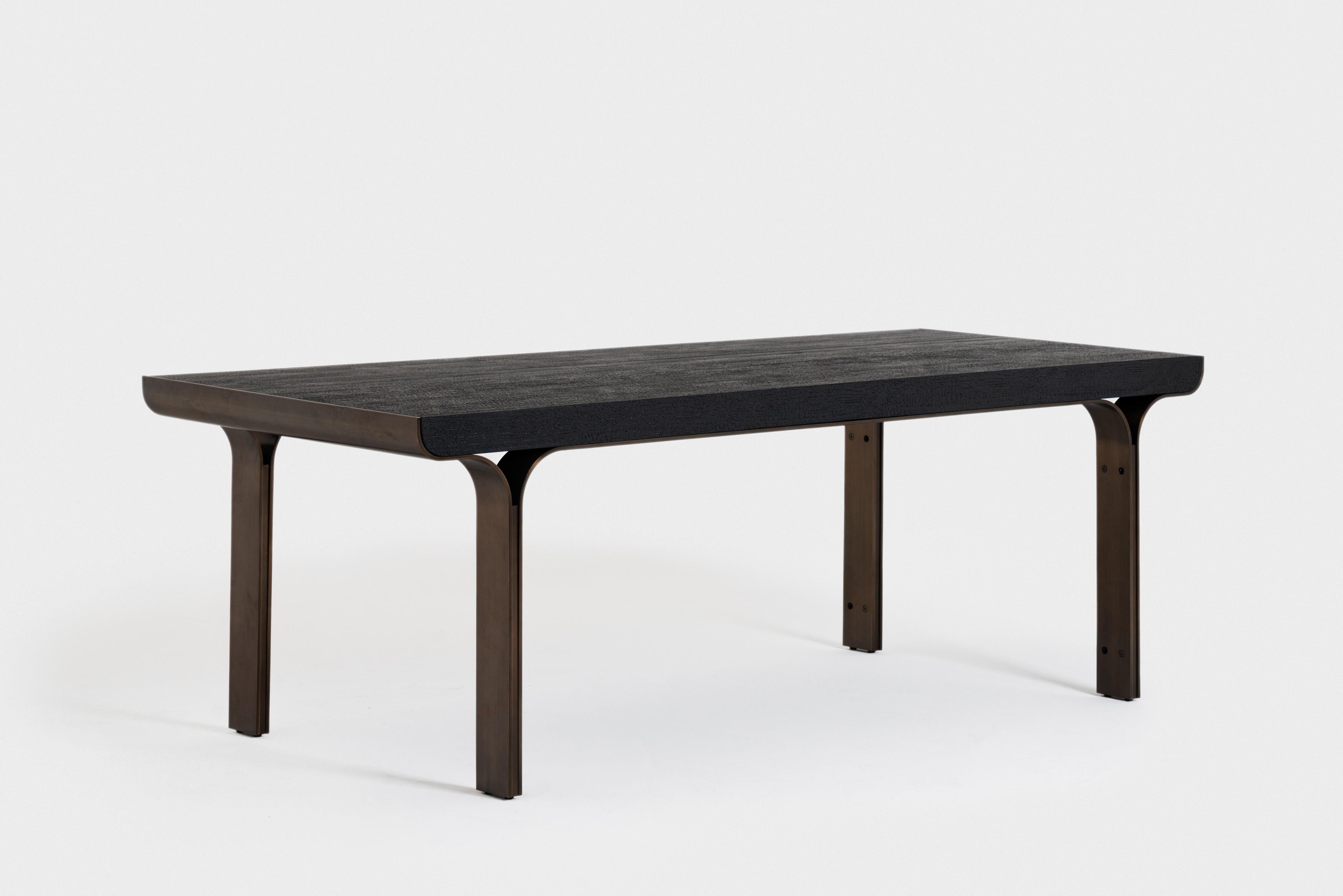La table de salle à manger Osvaldo est dotée d'une structure en fer bruni qui supporte un plateau en bois brûlé à la texture foncée. La lumière qui traverse les pieds bronzés projette des ombres chaudes et complexes tout en rebondissant sur la