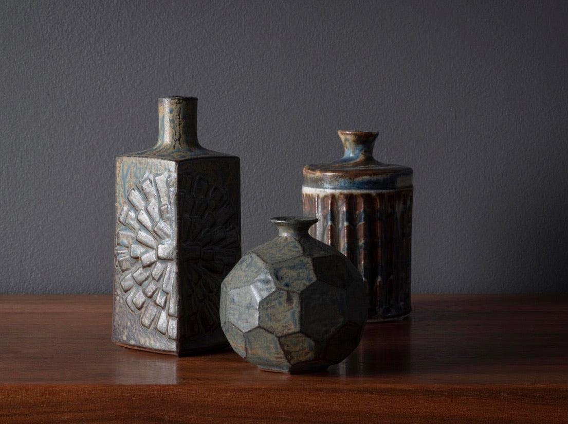 Collection of three Otagiri earth-toned vases with semi matte mottled glaze. Each piece features a unique decorative program; the tallest bottle has a raised starburst pattern, whereas the smaller vessels have carved and faceted geometric details.