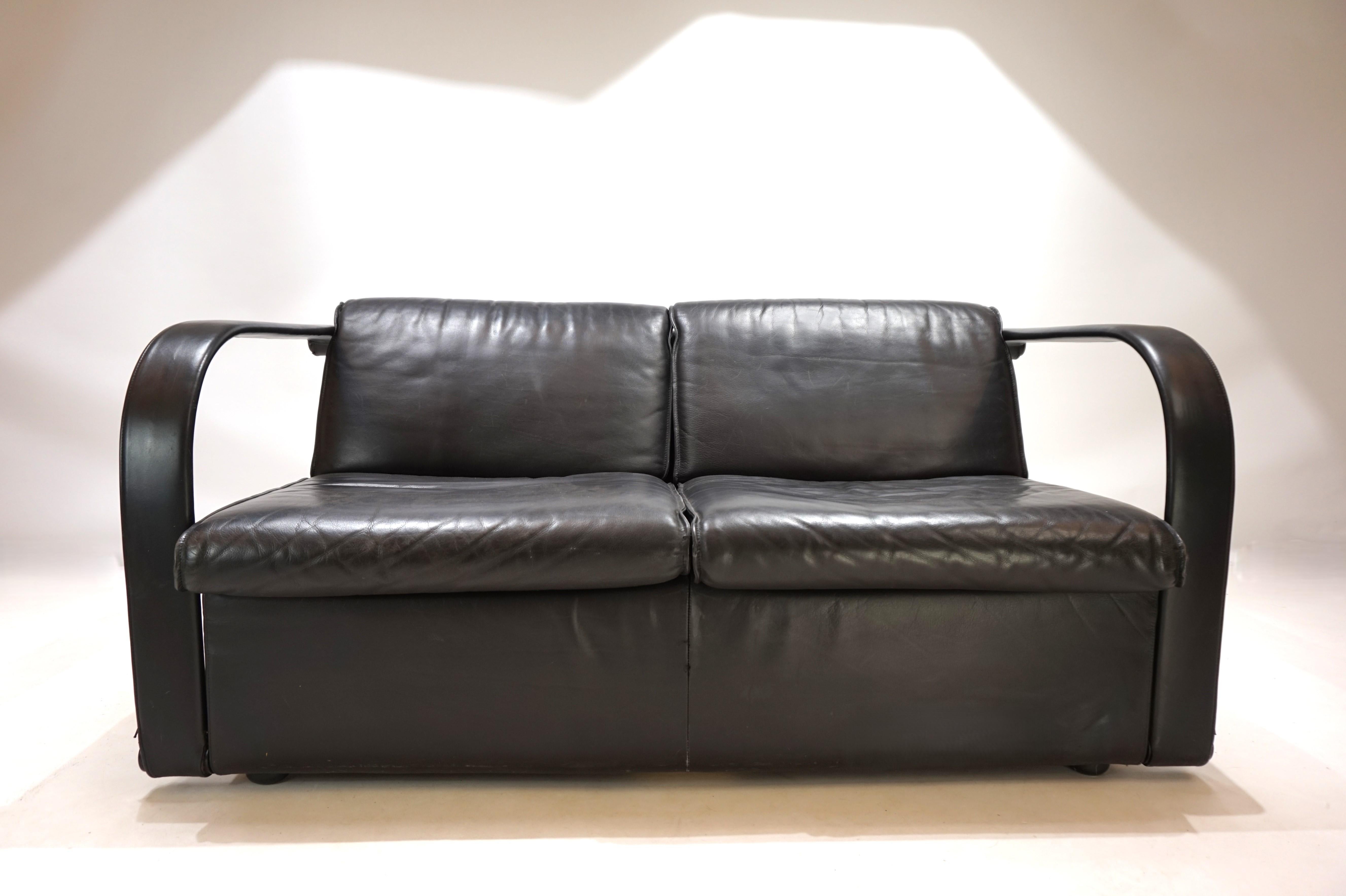 Questo divano in pelle Arconas è in ottime condizioni. La spessa pelle nera presenta minimi segni di usura. I cuscini dello schienale e della seduta, così come i rivestimenti in pelle dei braccioli, sono fissati con un sistema di cerniere e possono