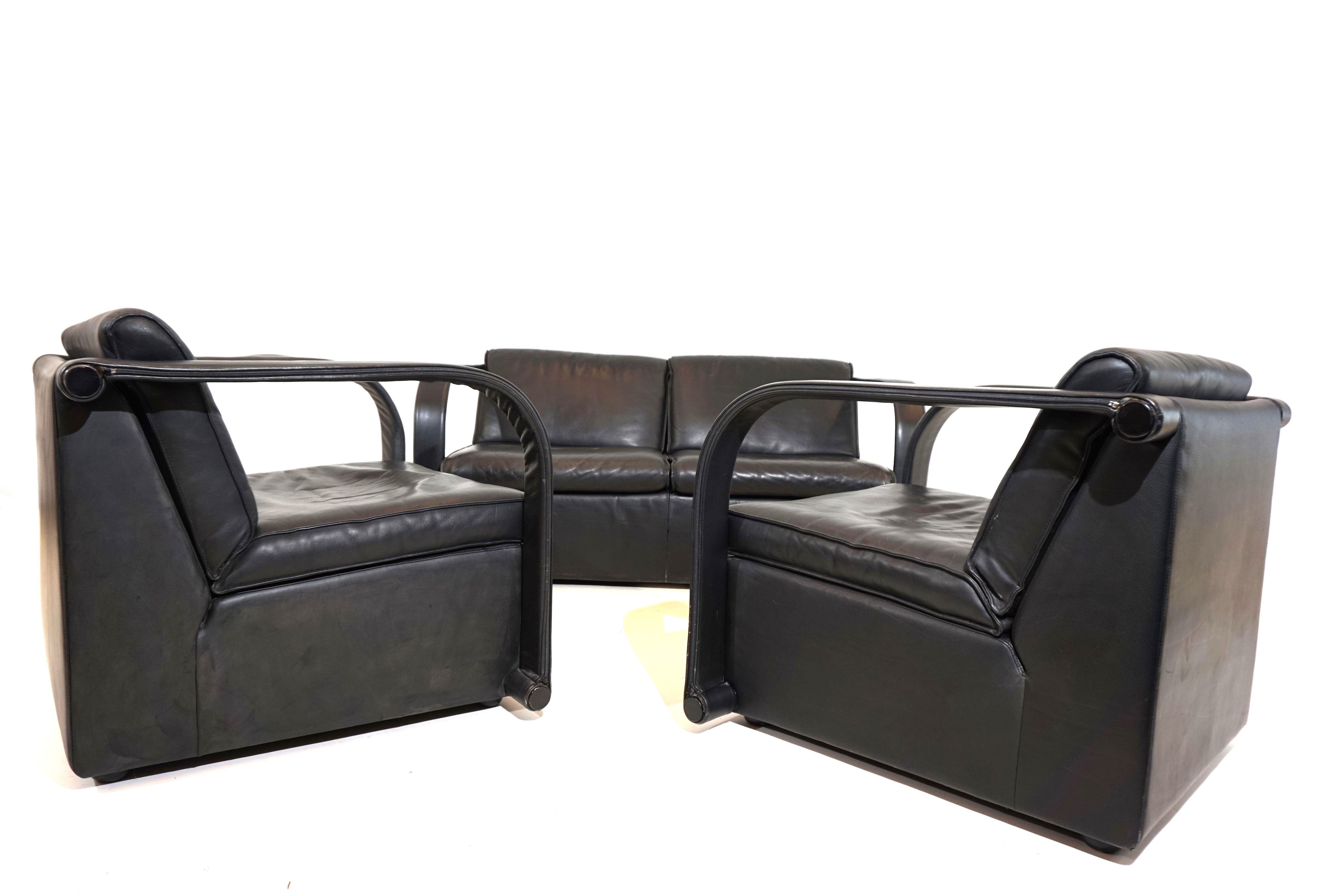 Die Ledergarnitur Arconas besteht aus einem 2-sitzigen Sofa und zwei Sesseln. Das dicke schwarze Leder ist in sehr gutem Zustand mit minimalen Gebrauchsspuren. Die Rücken- und Sitzkissen sowie die Lederbezüge an den Armlehnen sind mit einem