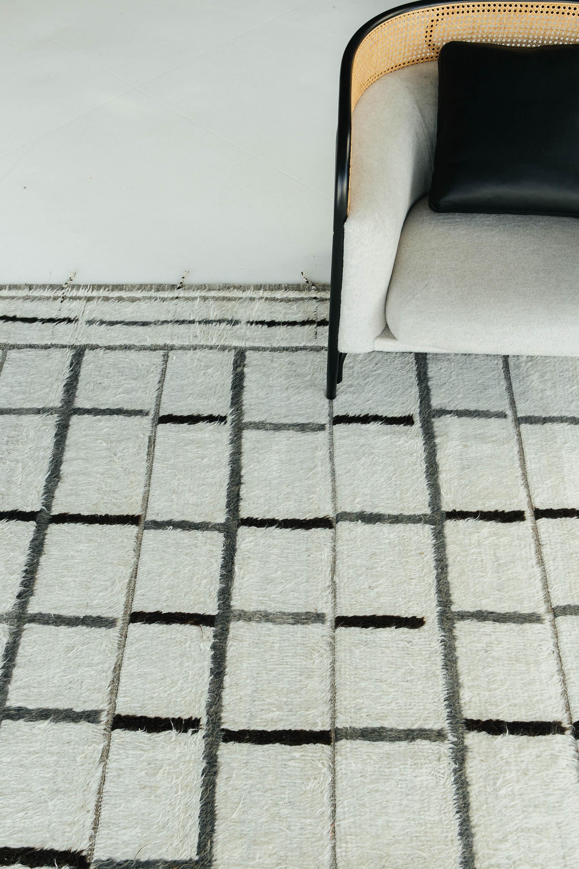 A white surface of handwoven wool designed with embossed line work with a grey and black checkered layout on top of a natural flat-weave. Designed in Los Angeles with inspiration from Moroccan tribal styles as a contemporary interpretation for the