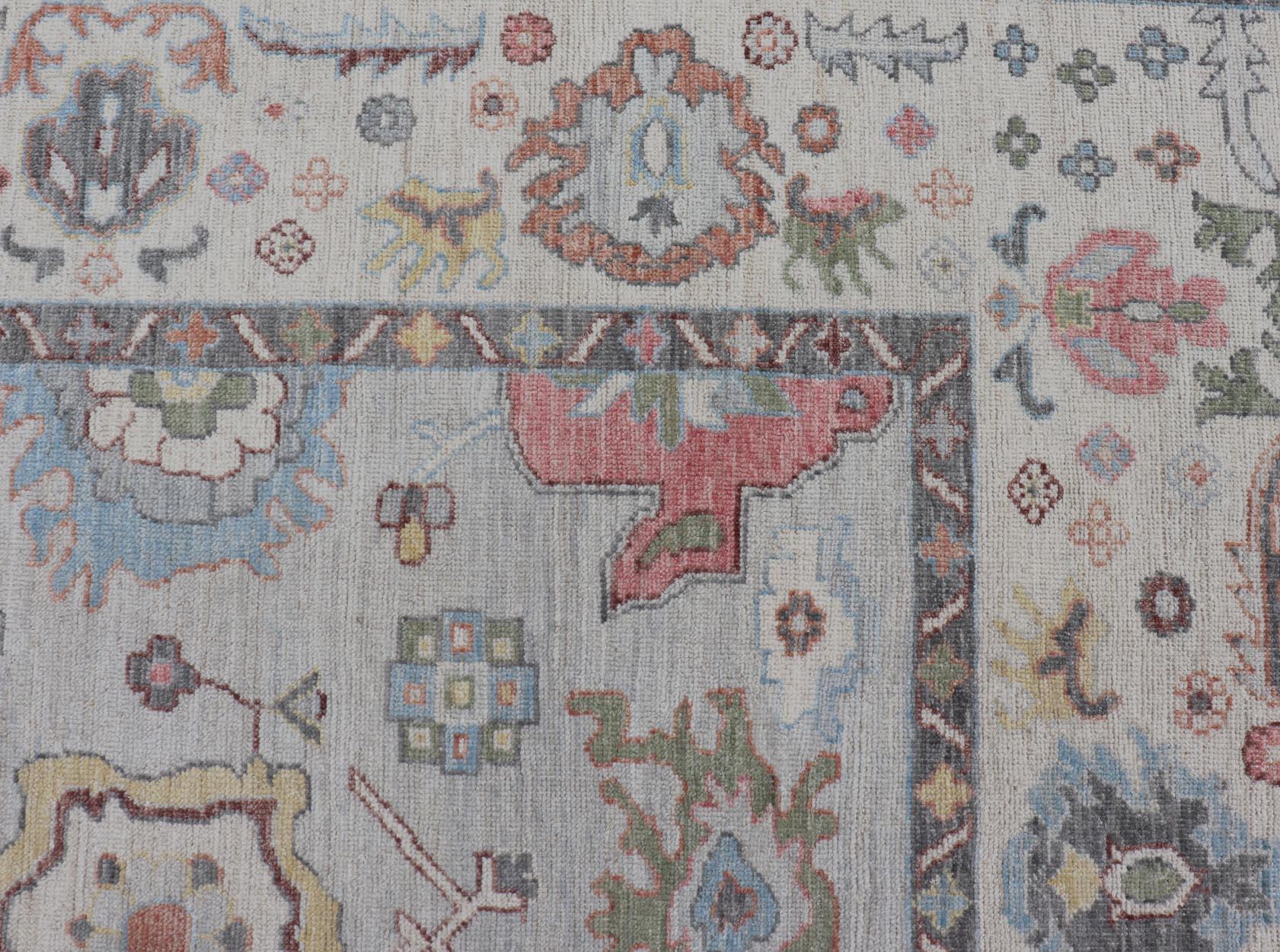 The border and field are covered in floral-like motifs filled with colors such as pink, green, gold, teal and red-oranges on top of silvery grey background. The border showcases a white-gray between two bands of a rich charcoal, edged with a small,