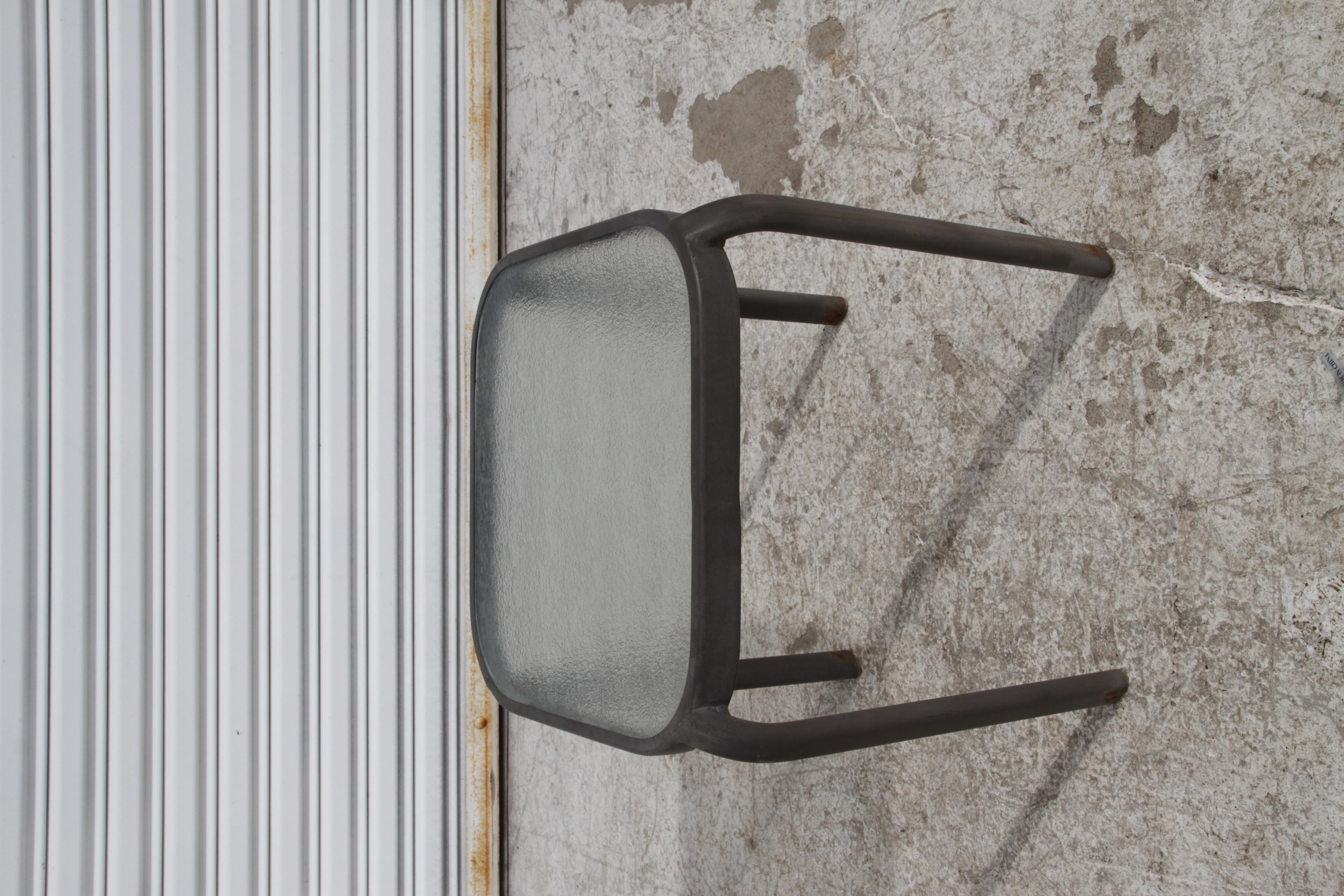 Dies ist ein Beistelltisch für den Außenbereich mit einem Metallgestell und einer Glasplatte. Das Gestell ist schwarz oder dunkelgrau und hat ein Rohrdesign mit geschwungenen Beinen, die aus der Tischplatte herausragen. Die Glasplatte scheint