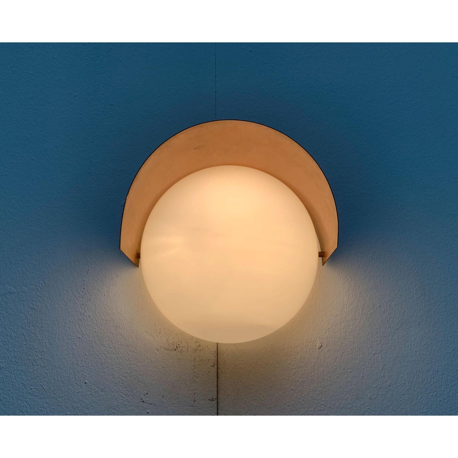 A wall lamp of traditional materiality and form. The diffuser is a hood made of mouth-blown opal glass. The body of the lamp with the screen is made of copper. The barrel-shaped shade sits above the glass. It is mounted with spacers to create a