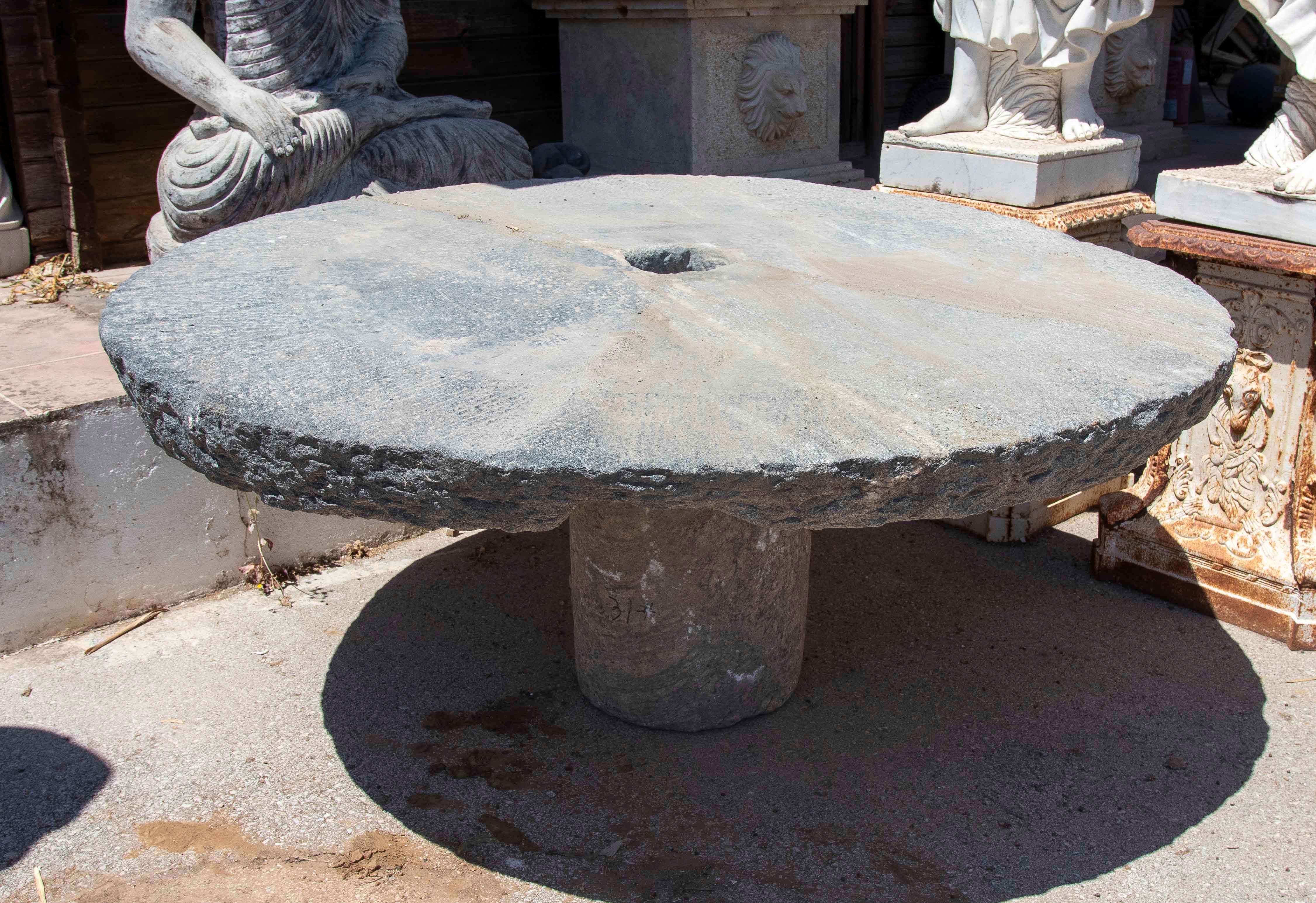 This unique table is crafted from an authentic antique millstone, repurposed and adapted as an outdoor tabletop. Its circular surface retains the original central opening, a functional characteristic of traditional millstones, which now provides a