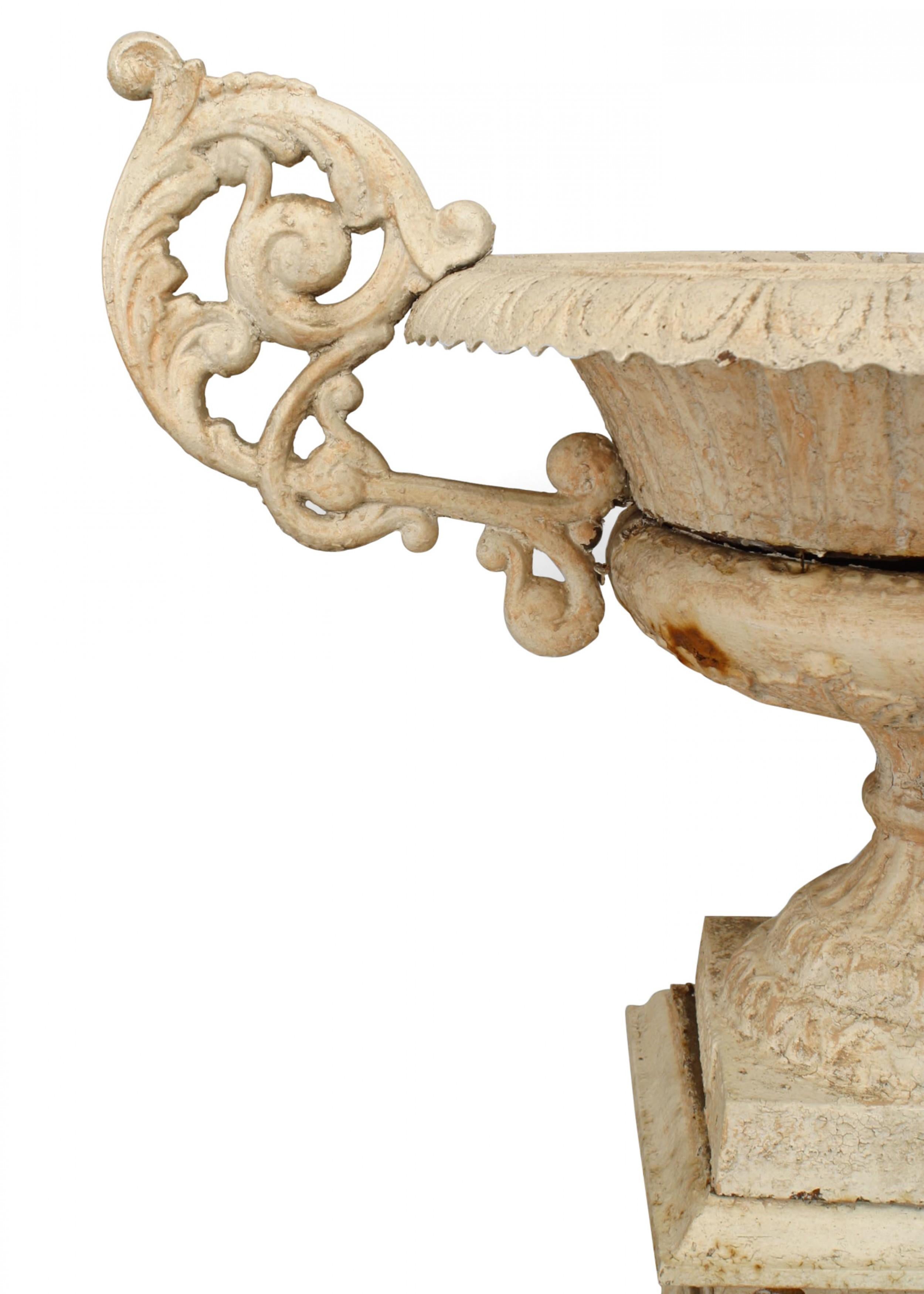 Outdoor American Victorian white painted round iron urn with large scroll handles and scalloped top Set on a square base.

One handle is damaged and has been wired in place. Finish is distressed.