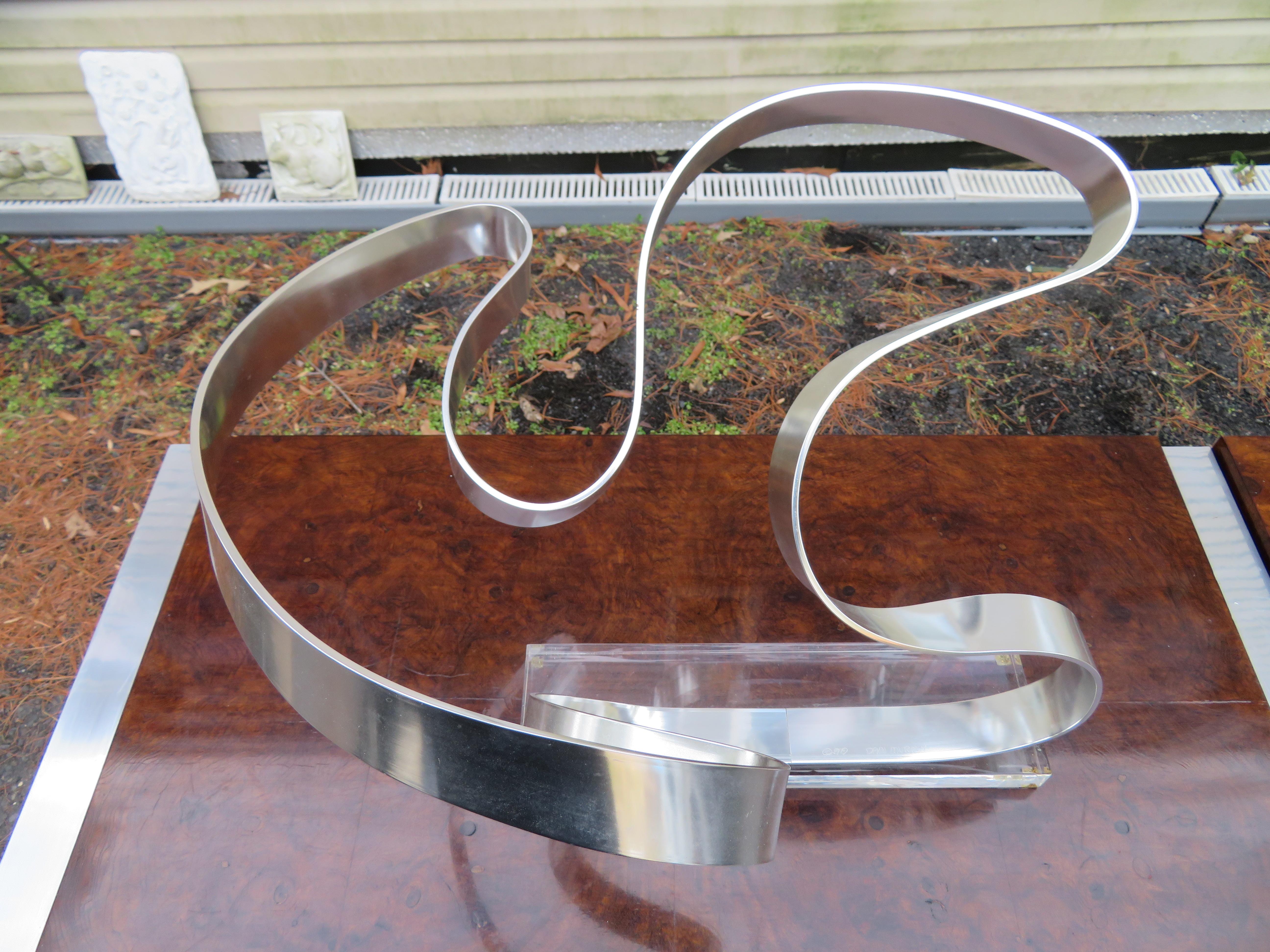 This fantastic sculpture was created by famed artist Dan Murphy in 1979. This abstract aluminum ribbon sculpture is mounted on a rectangular lucite base. This sculpture measures 13