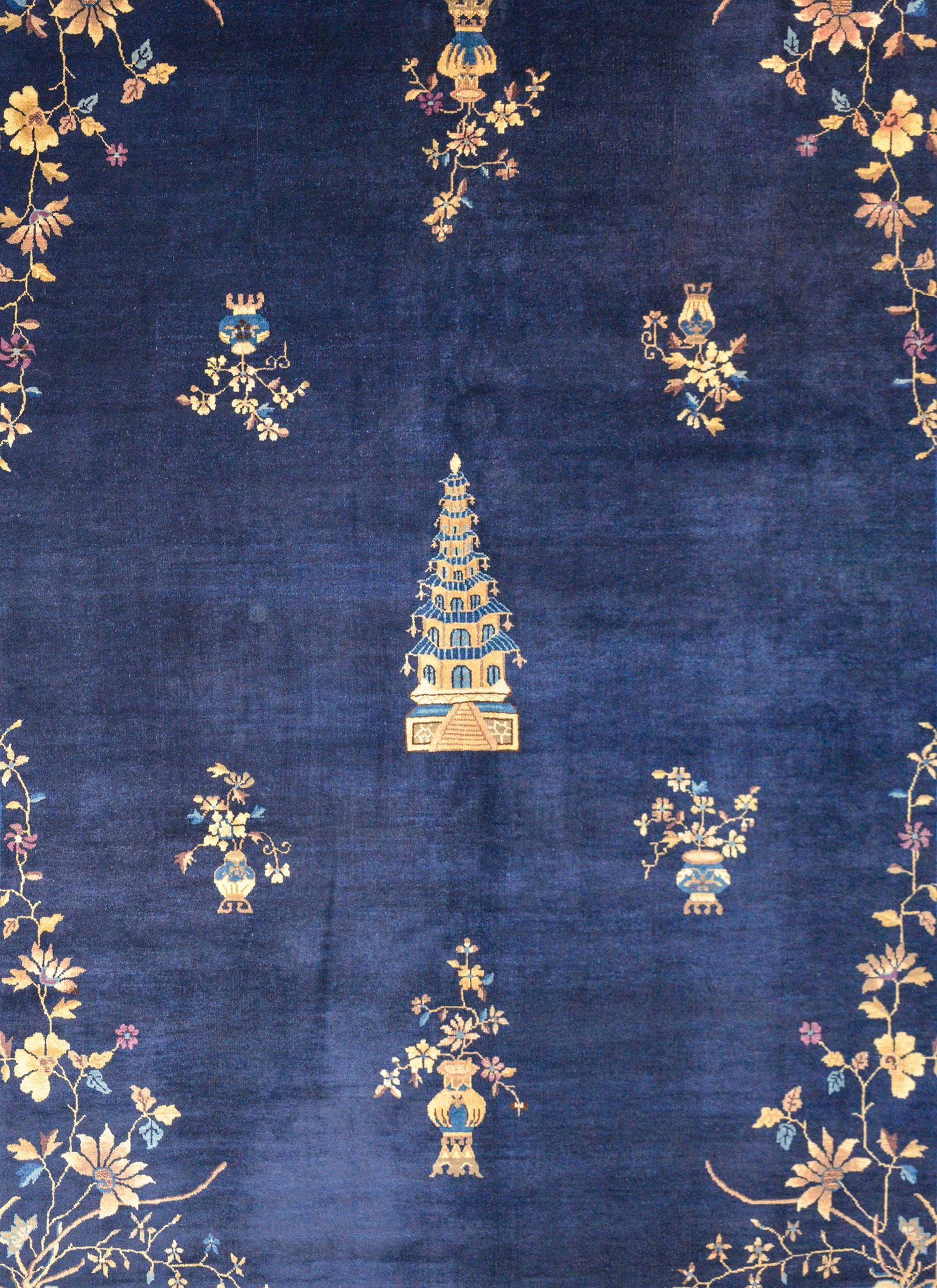 An outstanding early 20th century Chinese Art Deco rug with a large pagoda in the center amidst a field of potted auspicious flowers woven in cream, brown, light and dark indigo, against a dark indigo field and surrounded by a wide lighter indigo