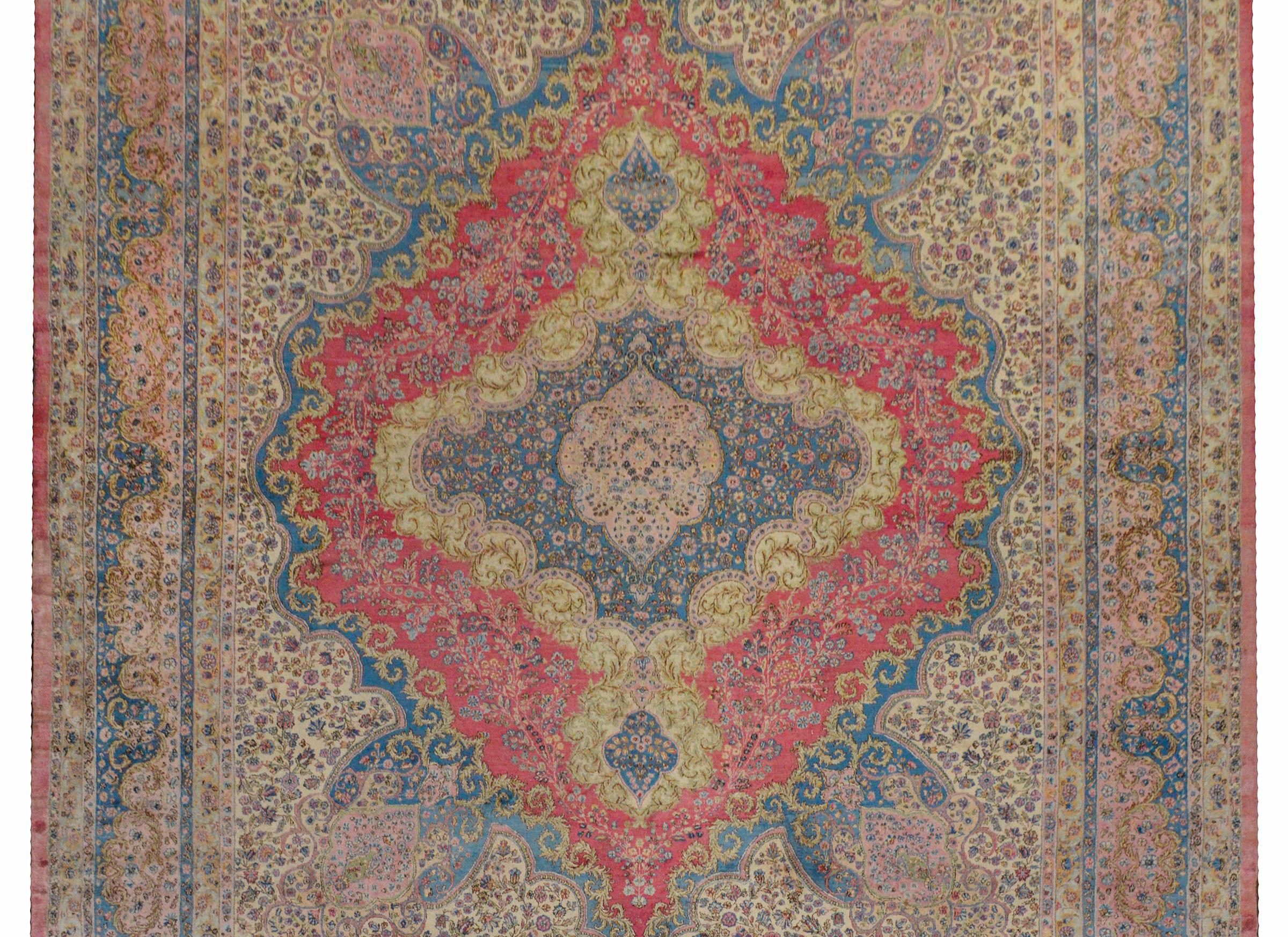 An outstanding early 20th century palatial Persian Kirman rug with an incredible large-scale floral diamond medallion woven in light indigo, gold, pale green, and pink amidst a field of flowers on a cream colored background. The border is Grande