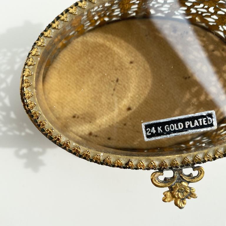Une façon fabuleuse d'exposer vos bijoux préférés. Cette boîte à bijoux ovale à pied est plaquée en or 24K. Il présente des côtés percés et un plateau en verre avec un couvercle à charnière. Le côté est orné d'un motif floral et la poignée de la