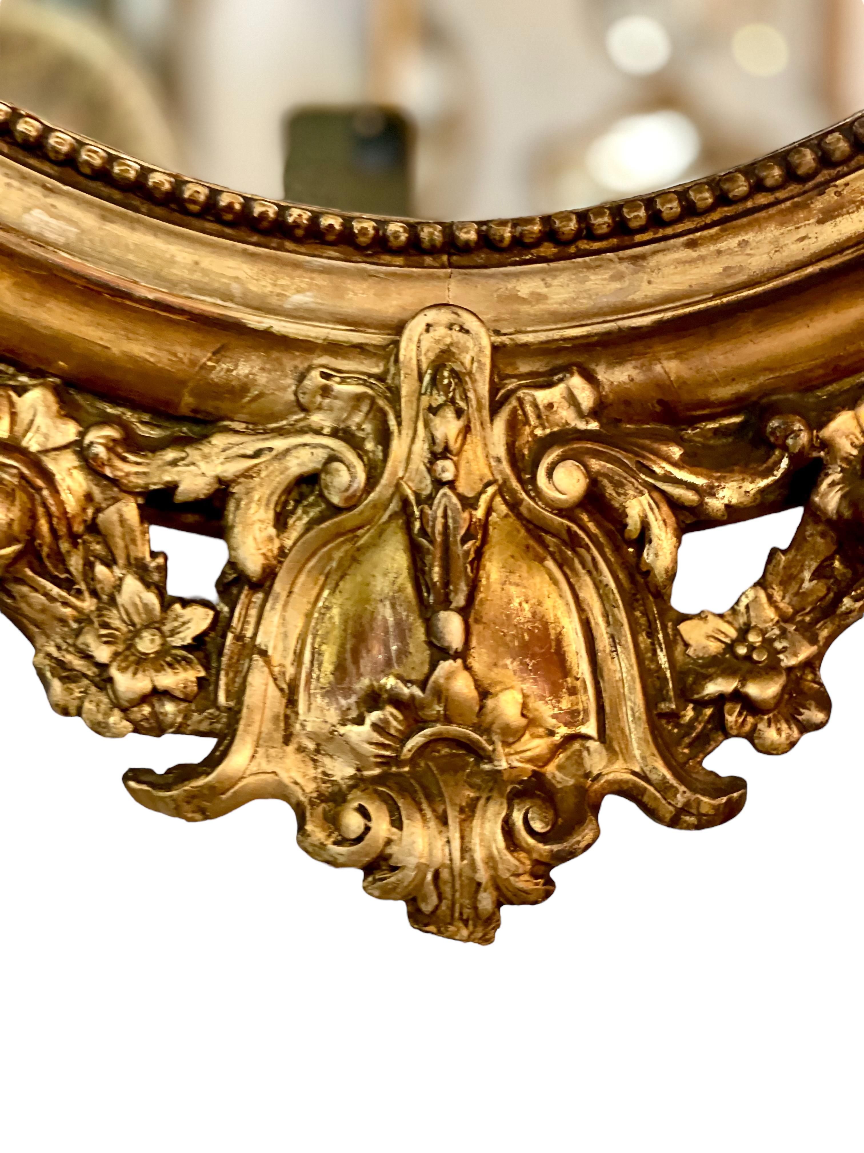 A very striking oval mirror in wood and gilded plaster, dating from the late 19th century. This stunning antique mirror features at its crest an intricate medallion-shaped pediment complete with miniature oval mirror, flanked on either side by