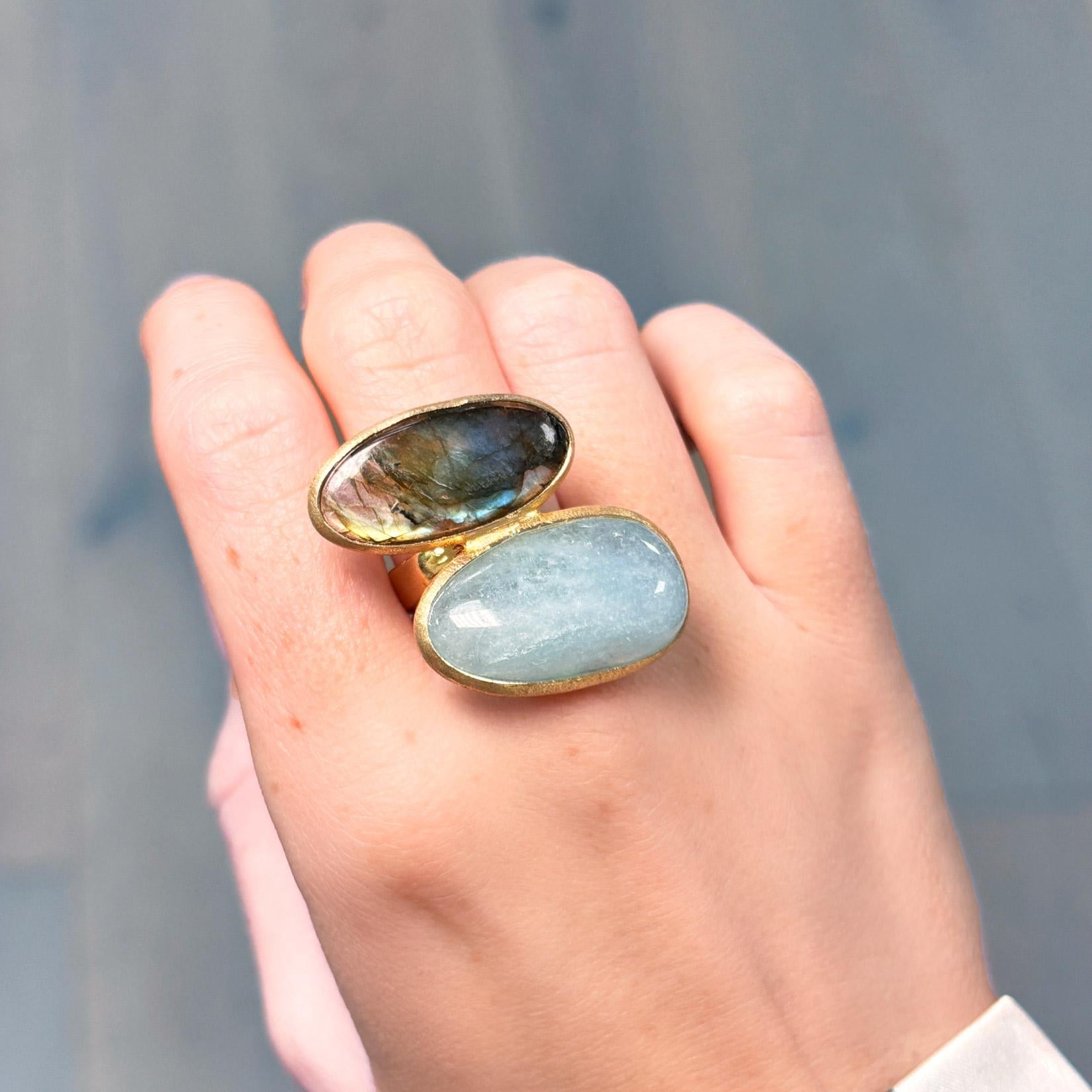 Scopri la magia del contrasto con questo squisito anello da cocktail a doppia pietra, caratterizzato dal sereno bagliore dell'acquamarina abbinato alla mistica iridescenza della labradorite. Ogni pietra dal taglio ovale è incastonata in un ricco