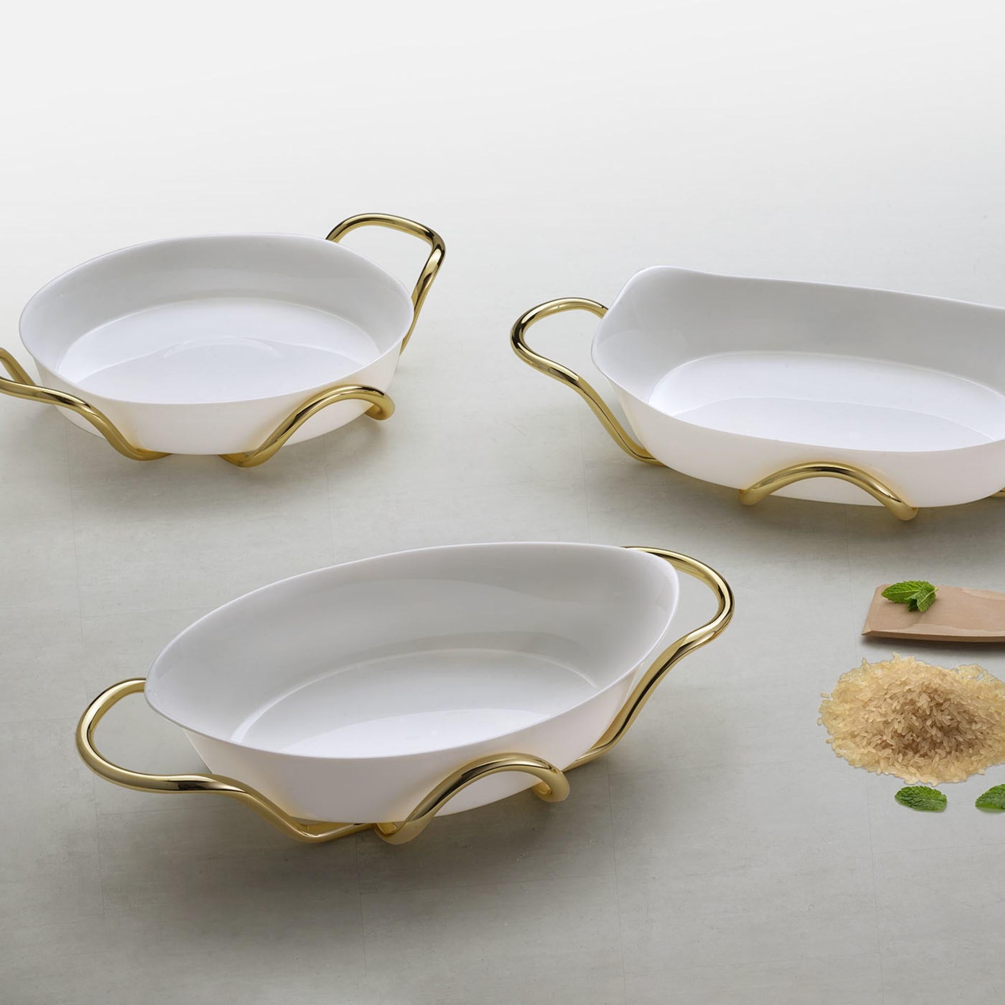 Plush and refined, this oval ceramic baking dish and its cylindrical sinuous-lined brass holder are superb for serving hot food with unprecedented elegance without direct contact with the table. A gleaming golden finish enriches the structure, which