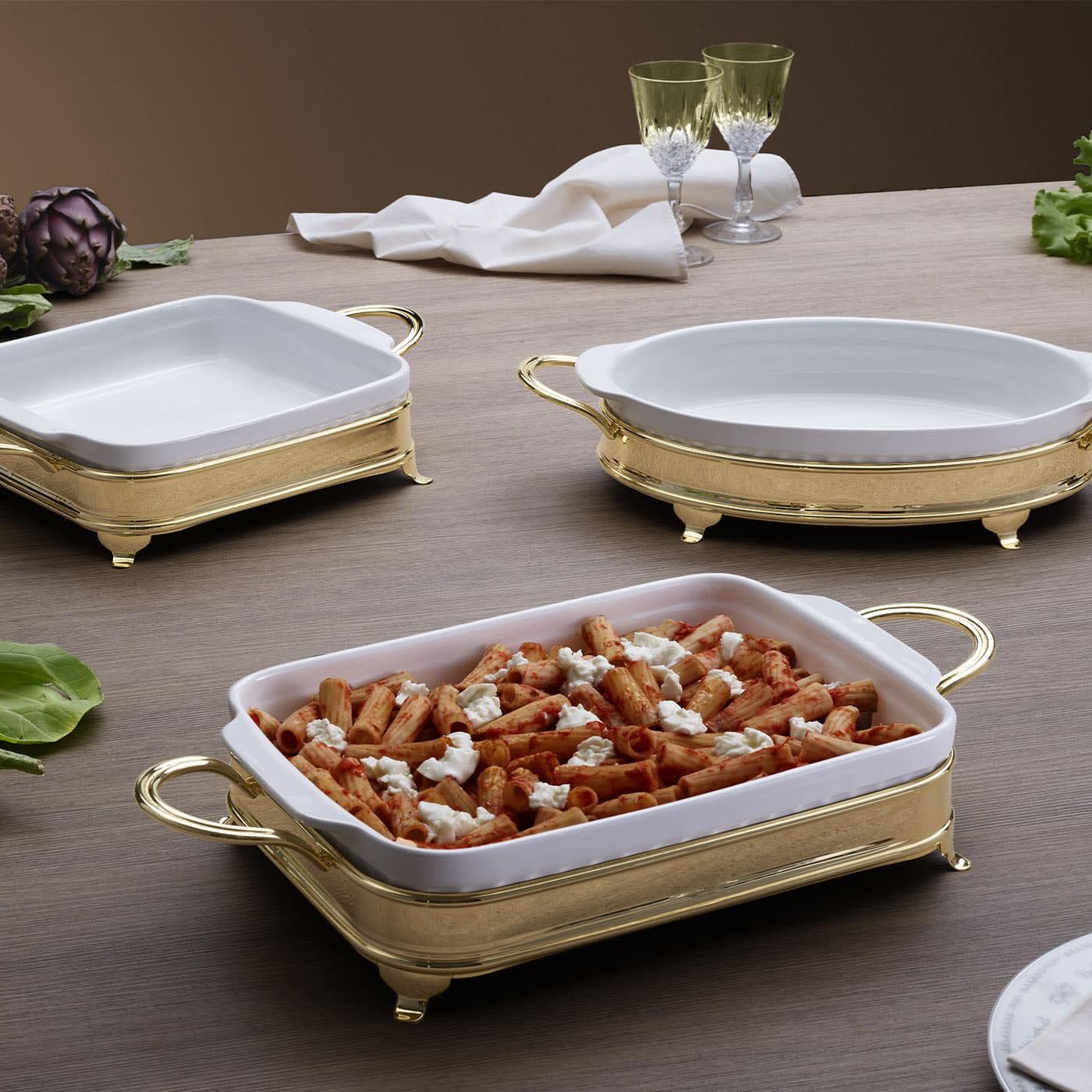 Designed for safely bringing hot food immediately to the table without renouncing elegance, this set comprises an oval baking dish in fine white ceramic and a luxurious golden-finished brass holder. Its tiny feet allow for avoiding contact with the
