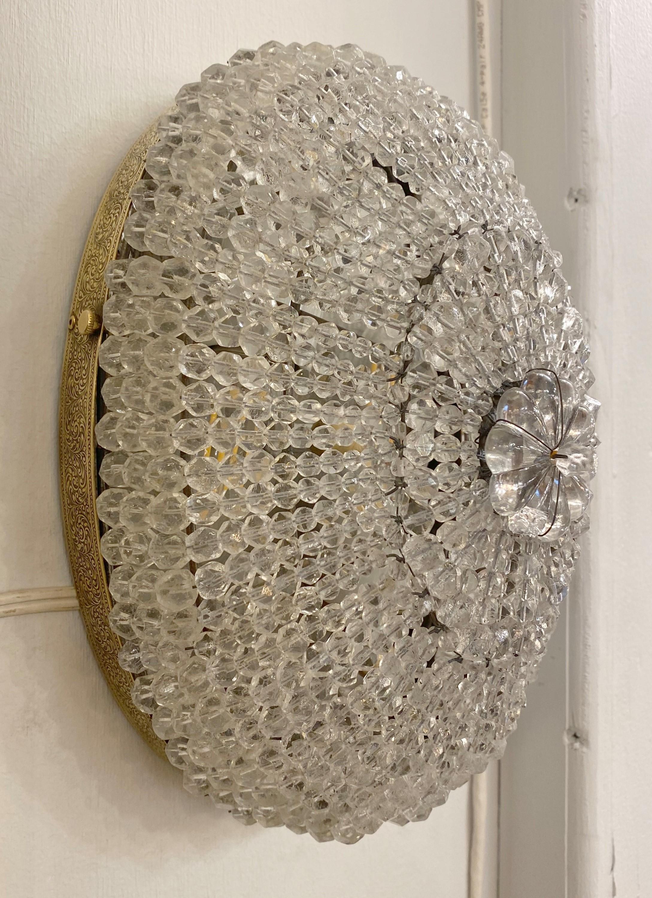Early 20th Century oval wall sconce featuring clear beaded strands of crystals and an ornate brass flush mount frame with swirl details. This sconce also has a switch installed on it's side to be individually turned off and on. It can also be used