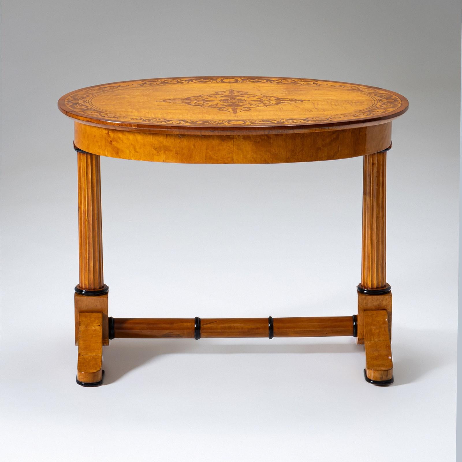 An elegant Biedermeier center table crafted from cherrywood, circa 1840. The table features an oval tabletop with a smooth apron and rests on forked legs with fluted columns and ebonized profiles. The legs are connected by a circular stretcher