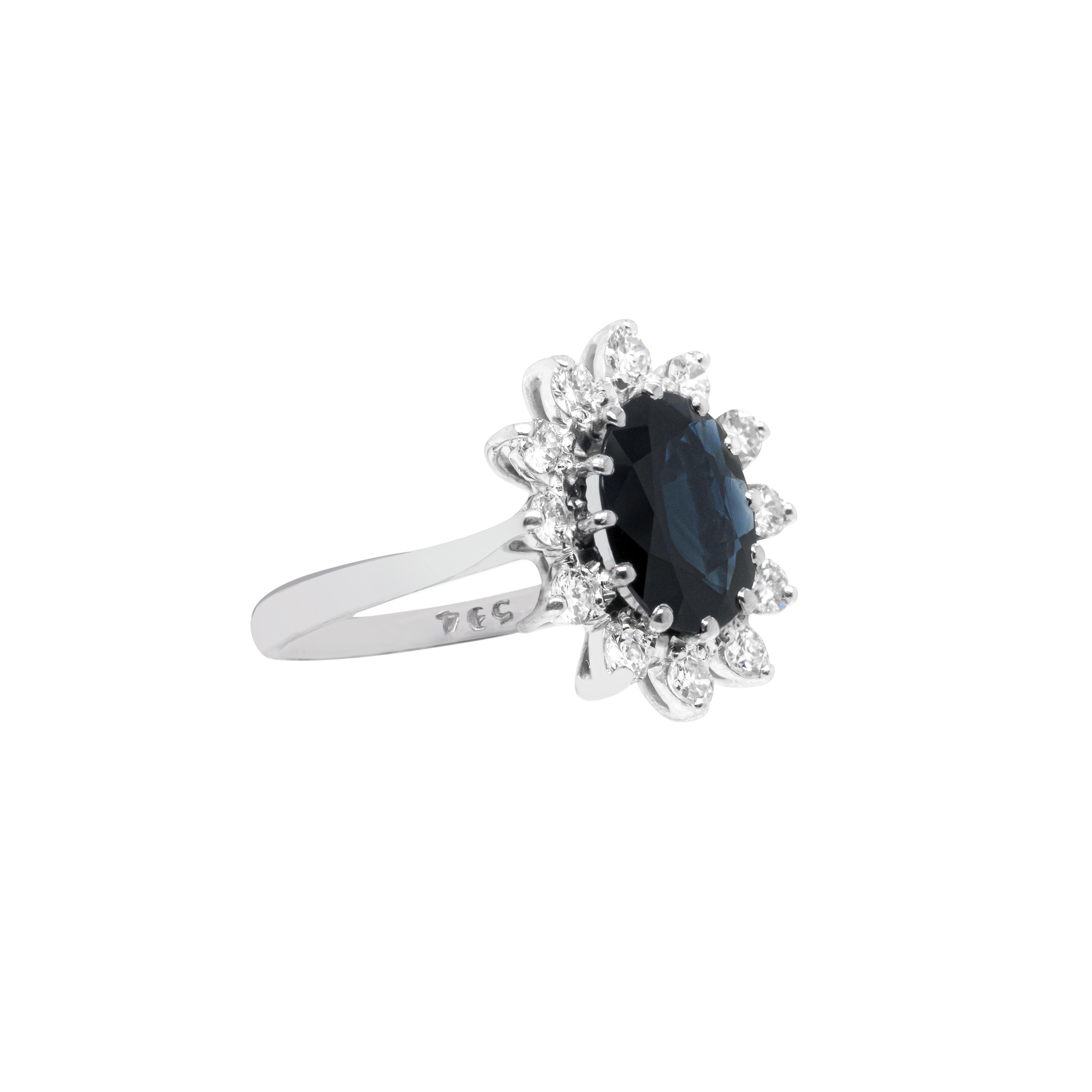 Reminiscent of the Princess Diana cluster engagement ring, this beautiful piece features a dark blue oval sapphire in the centre weighing approximately 2.50 carats mounted in a 12 claw, open back setting. The sapphire is wonderfully surrounded by 12