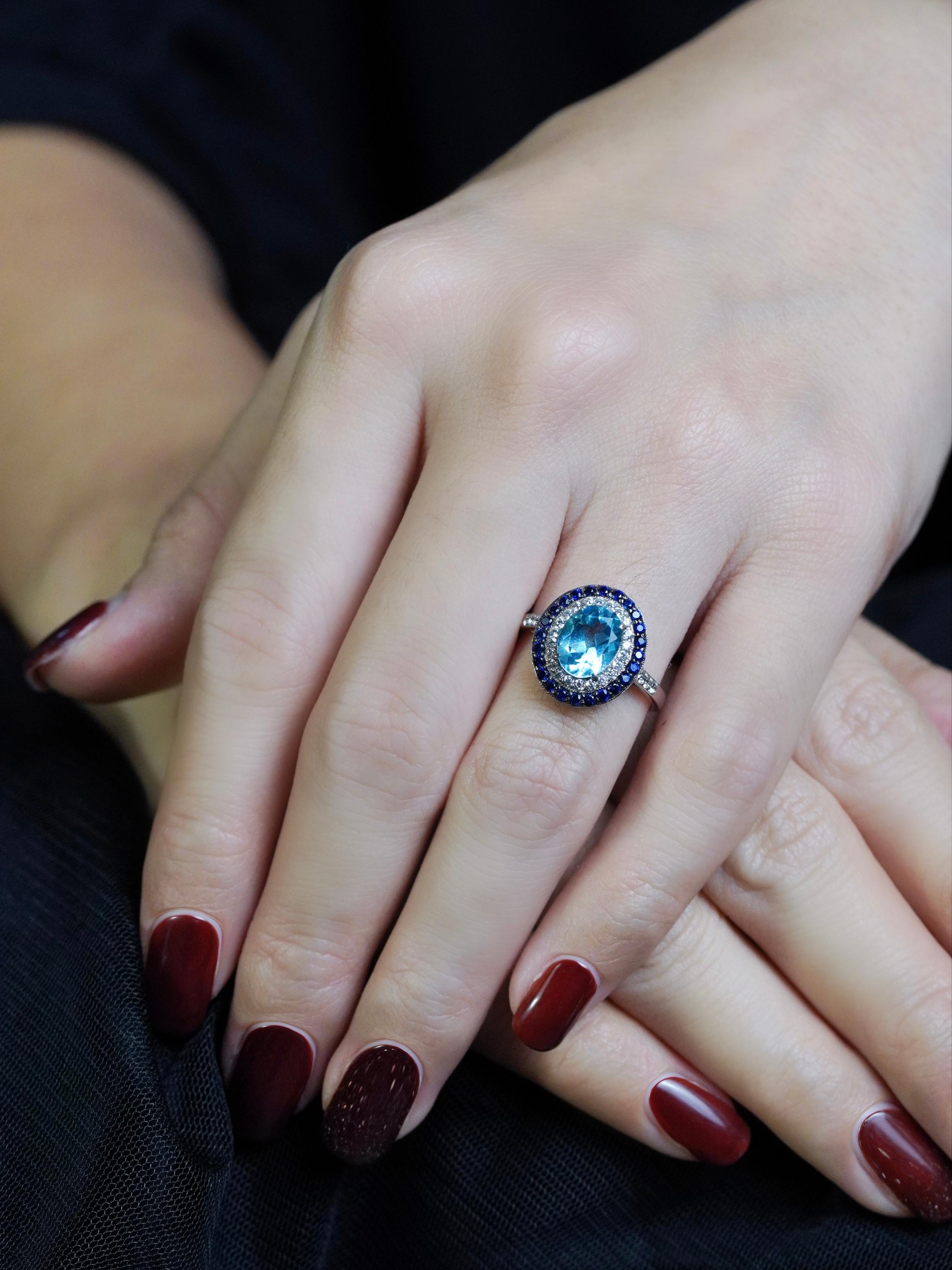 Questo anello in stile Art Déco, che emana eleganza a ogni sguardo, è una sinfonia di colori e scintille. Al centro, un topazio blu ovale brilla con serena brillantezza, evocando la calma di cieli limpidi e acque profonde. Circondato da un'aureola