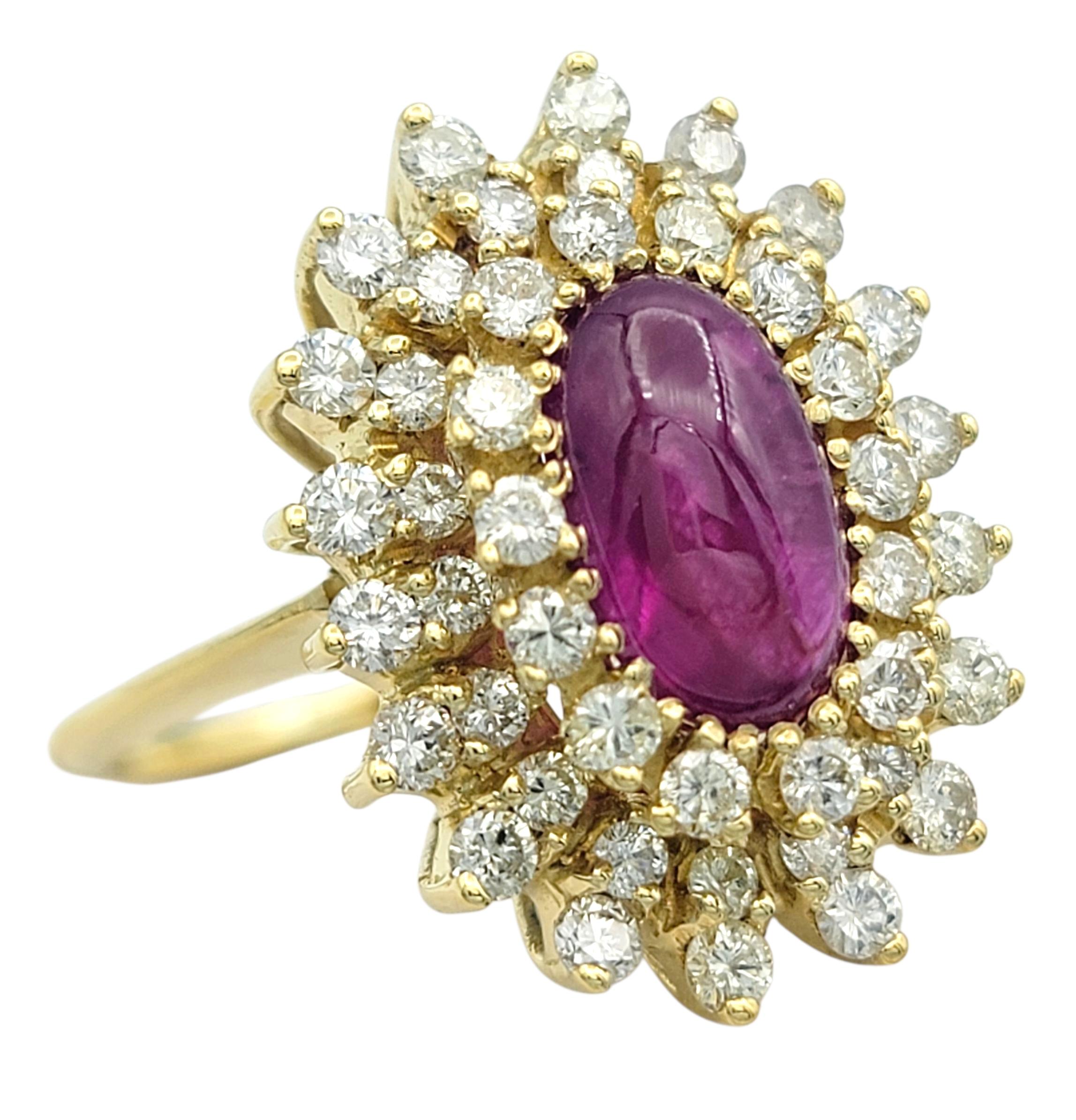 Cette étonnante bague en rubis et diamants est réalisée en or jaune 18 carats et présente en son centre un rubis cabochon ovale. La couleur rouge profond du rubis est encadrée par un double halo de diamants de taille ronde, créant un doux