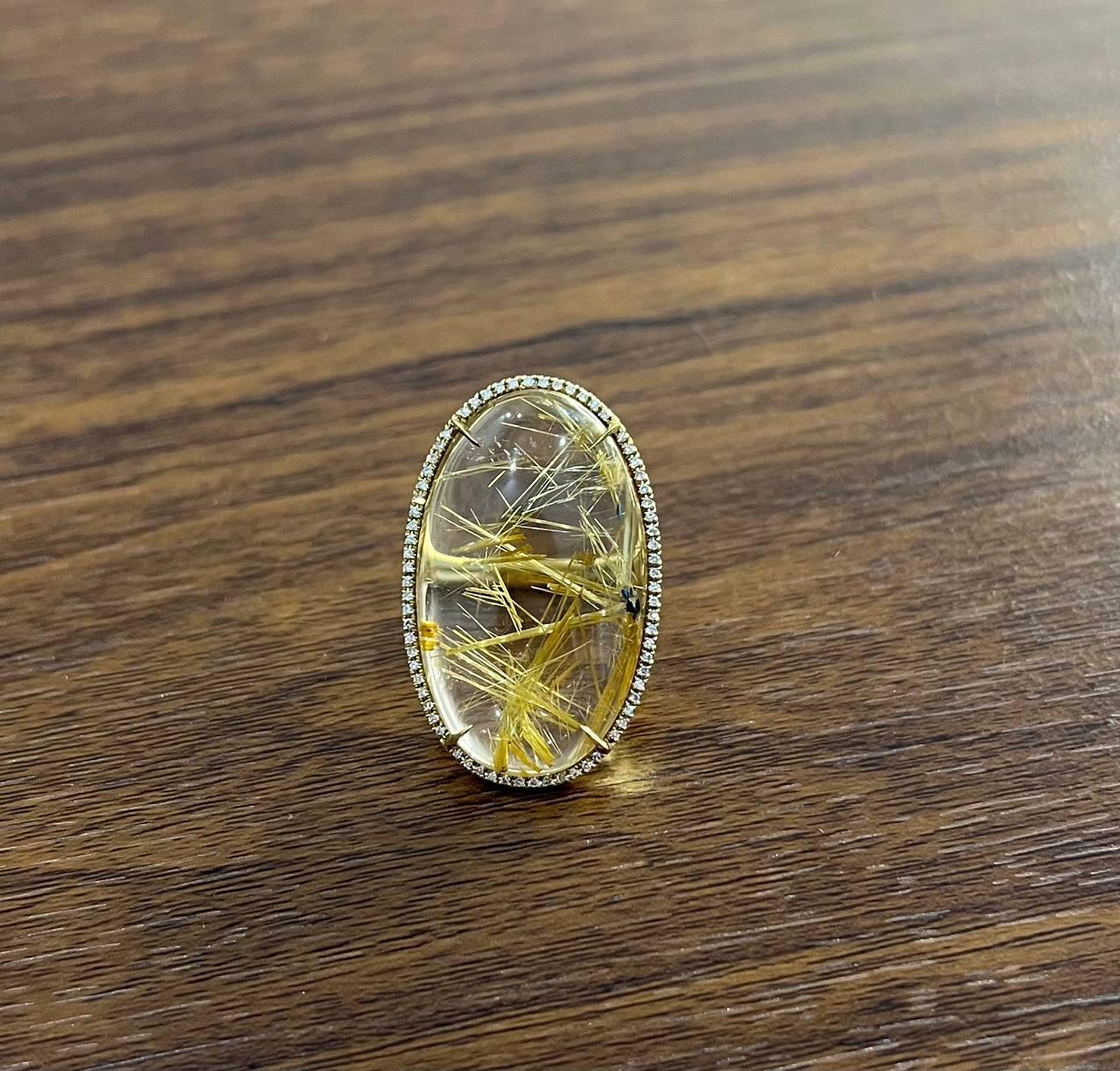 This striking ring features a large oval cabochon of rutilated quartz, showcasing golden needle-like inclusions. The quartz is set in an 18k gold bezel, surrounded by a delicate border of small diamonds. The intricate design highlights the natural