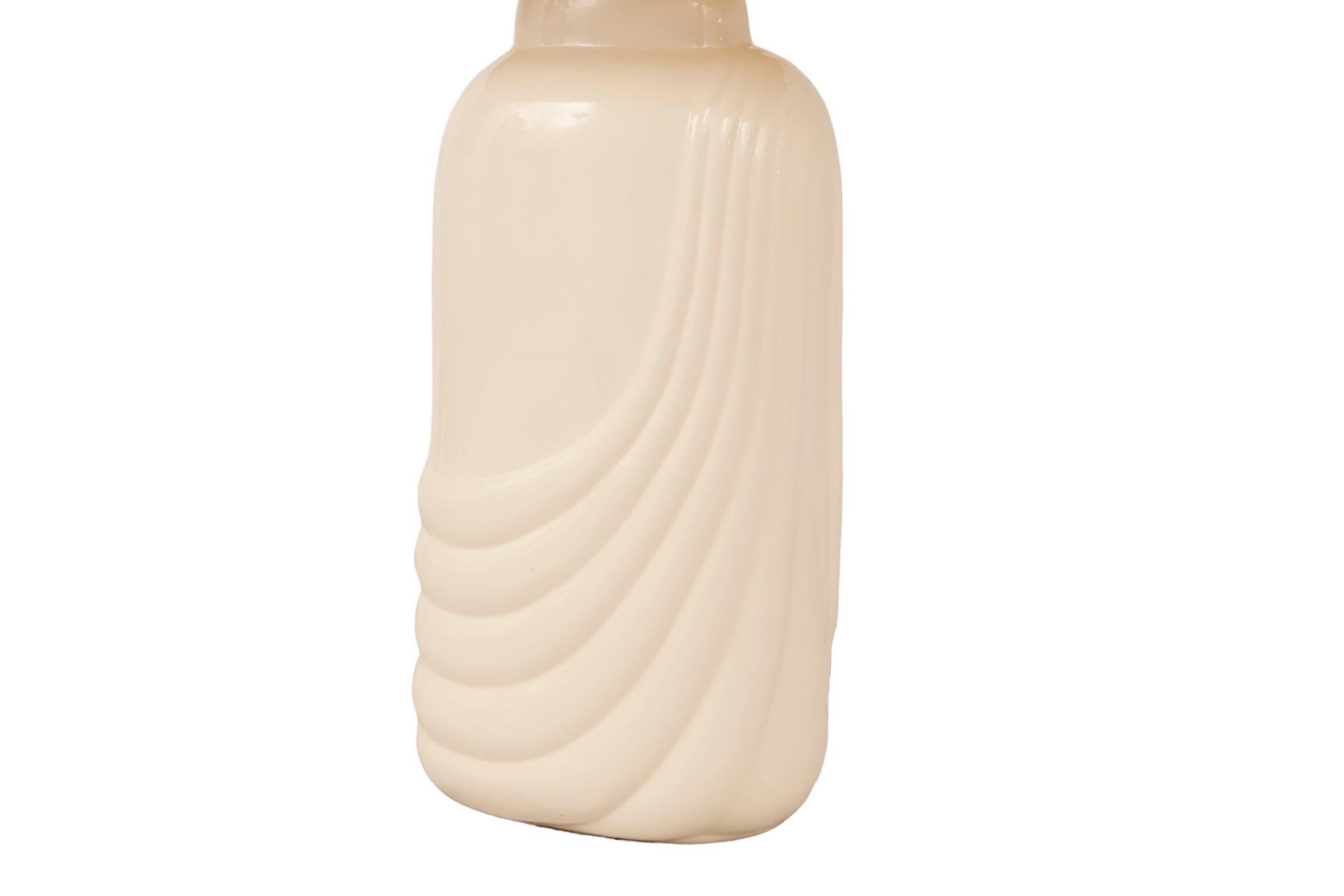 Lámpara ovalada de cerámica crema con pantalla a juego. Sobre un arpa, en una pequeña columna de latón, se asienta una sombra de rocalla. El cuerpo de cerámica tiene seis ondas dispuestas en forma de guirnalda desde el hombro derecho hacia el lado