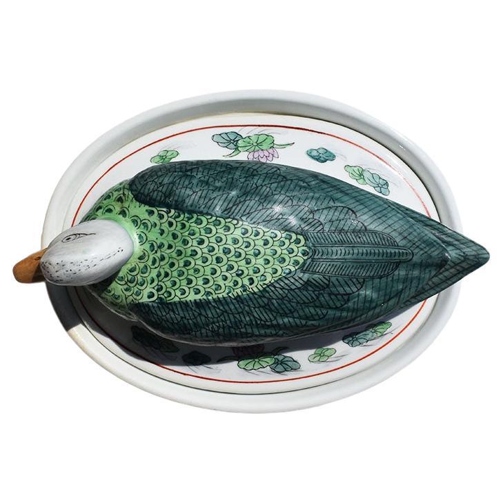 An oval ceramic duck motif tureen with a floral motif. Crisp white ceramic is hand-painted with a motif of lotus and lush green leaves in pink and blue. At the top, the lid is topped with a ceramic duck or mallard painted in a beautiful green
