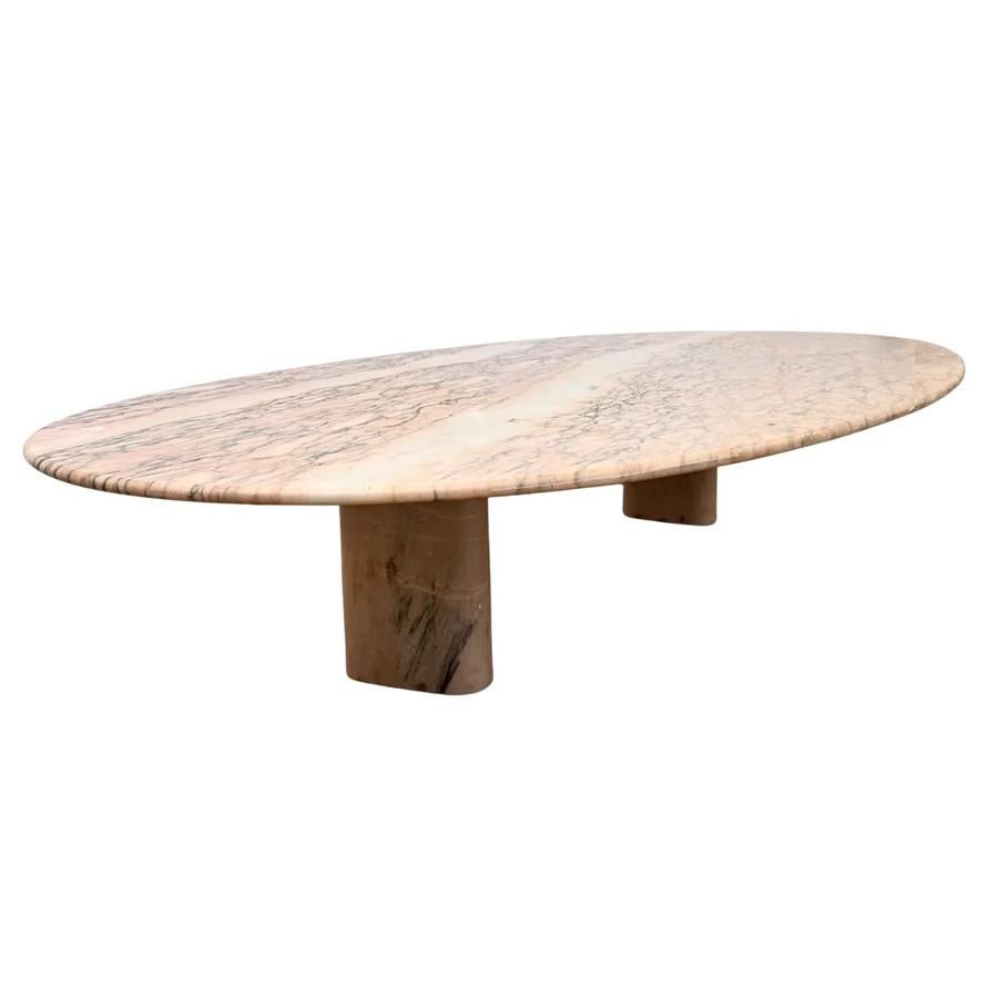 Oval marble coffee table in Rosa Aurora, a Portuguese limestone known for its soft pink base and delicate taupe veining. The thick slab top sits on two cylindrical legs in matching stone. Rounded edges and a minimalist silhouette keep the focus on