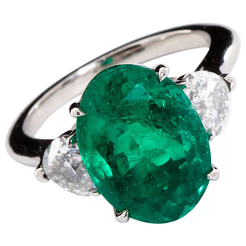 An unusual deep green 5.60 ct. oval modified brilliant cut natural Colombian emerald set between two heart shaped diamonds (1.01 ctw) in a platinum ring. The diamonds are F color, VS clarity. A superb emerald ring with the diamond hearts pushing