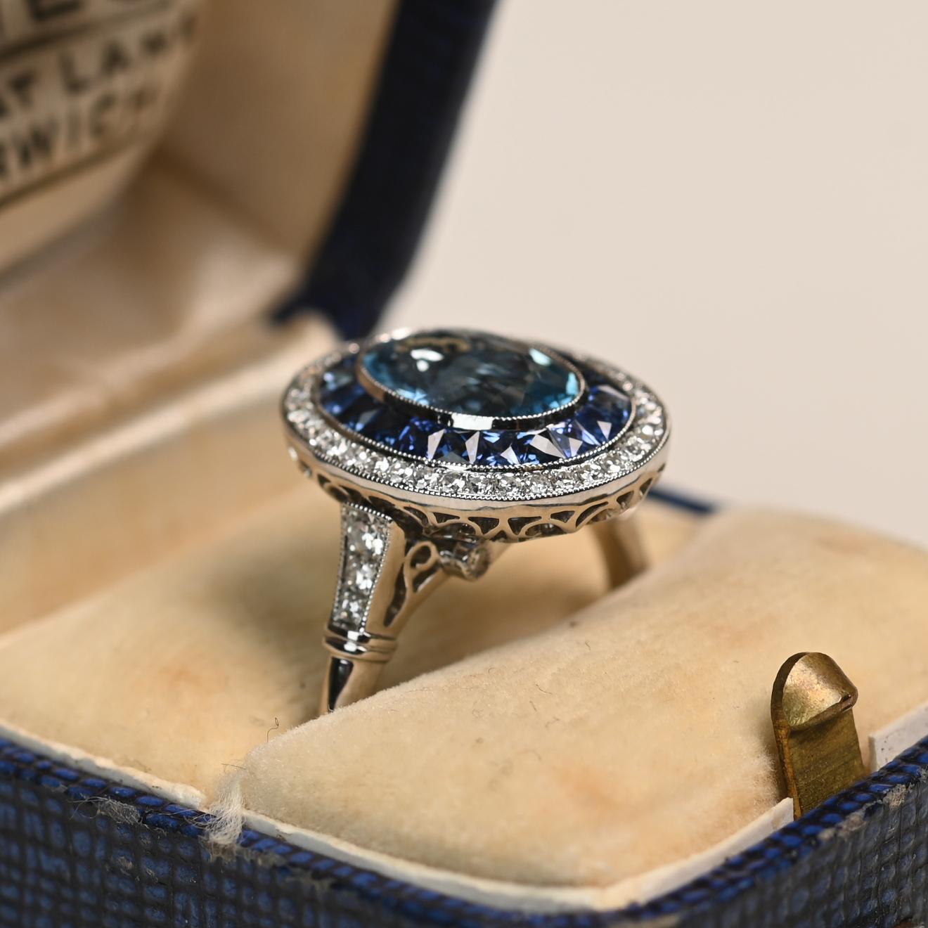 Cette bague exquise est ornée d'une aigue-marine de taille ovale d'environ 1,20 carat, d'une couleur Santa Maria très prisée. La pierre centrale vibrante est entourée d'un double halo saisissant de saphirs naturels et de diamants étincelants, créant