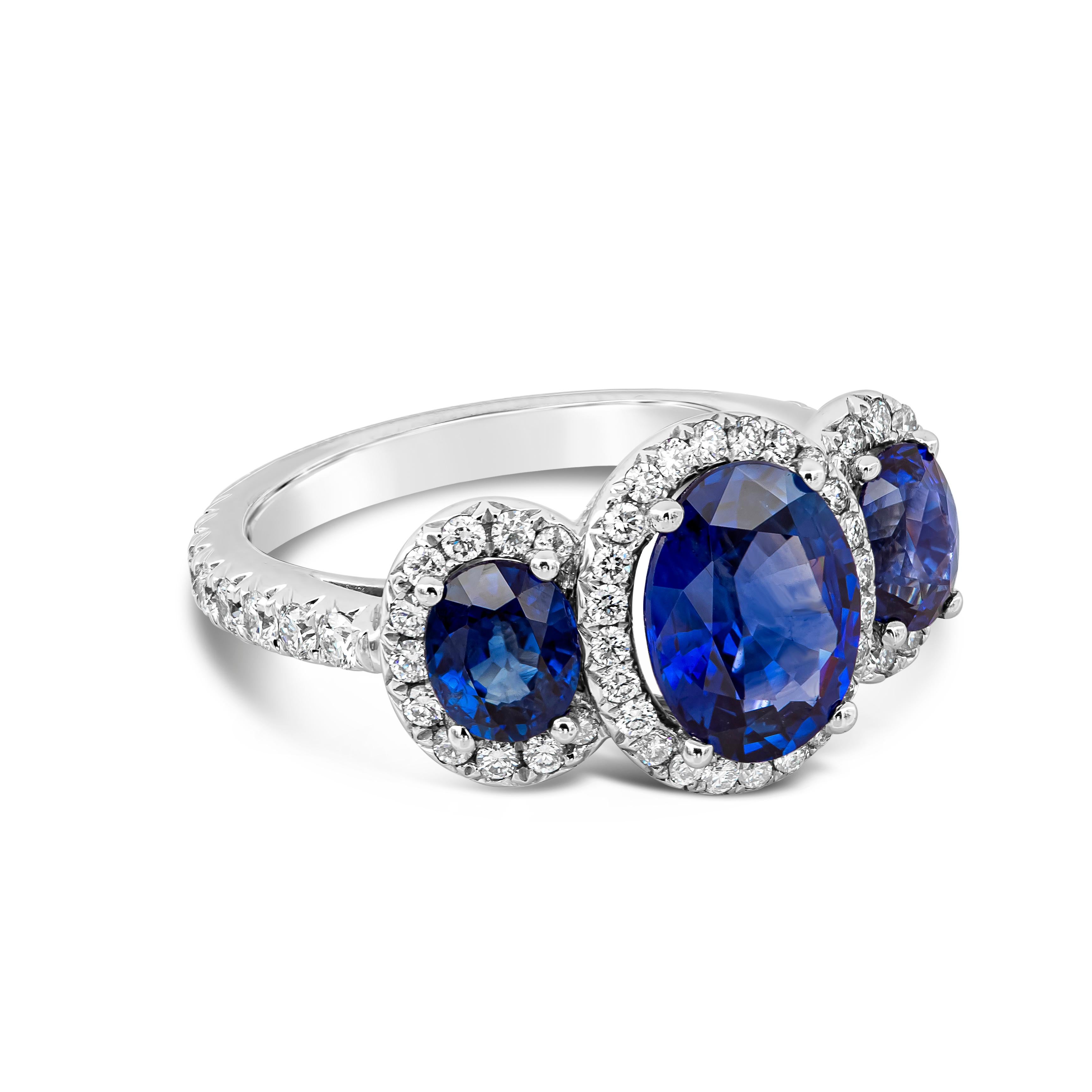 A vibrant and unique three-stone engagement ring style showcasing a 2.11 carat blue sapphire, flanked by smaller blue sapphires weighing 1.40 carats total on either side. Each stone is set in a brilliant diamond halo, set in a diamond encrusted