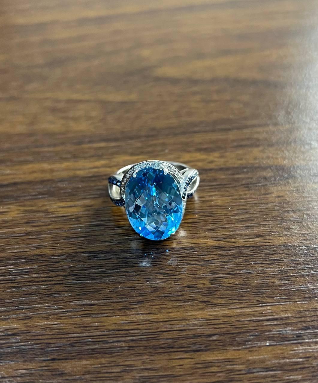 A striking and elegant ring featuring a large oval-shaped blue topaz with intertwining halo of diamonds and blue sapphires. The ring is crafted with intricate detailing in 14K White Gold