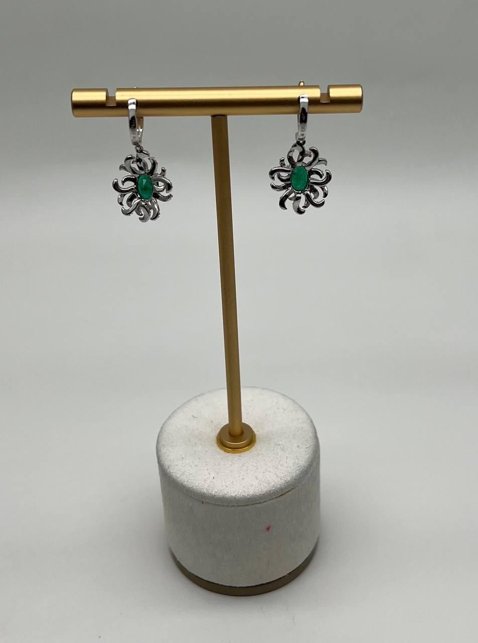 These elegant earrings feature vibrant green emeralds set in intricate, swirling designs. The gemstones are surrounded by delicate, sparkling diamonds, creating a captivating floral motif. The earrings are suspended from sleek, polished hoops,