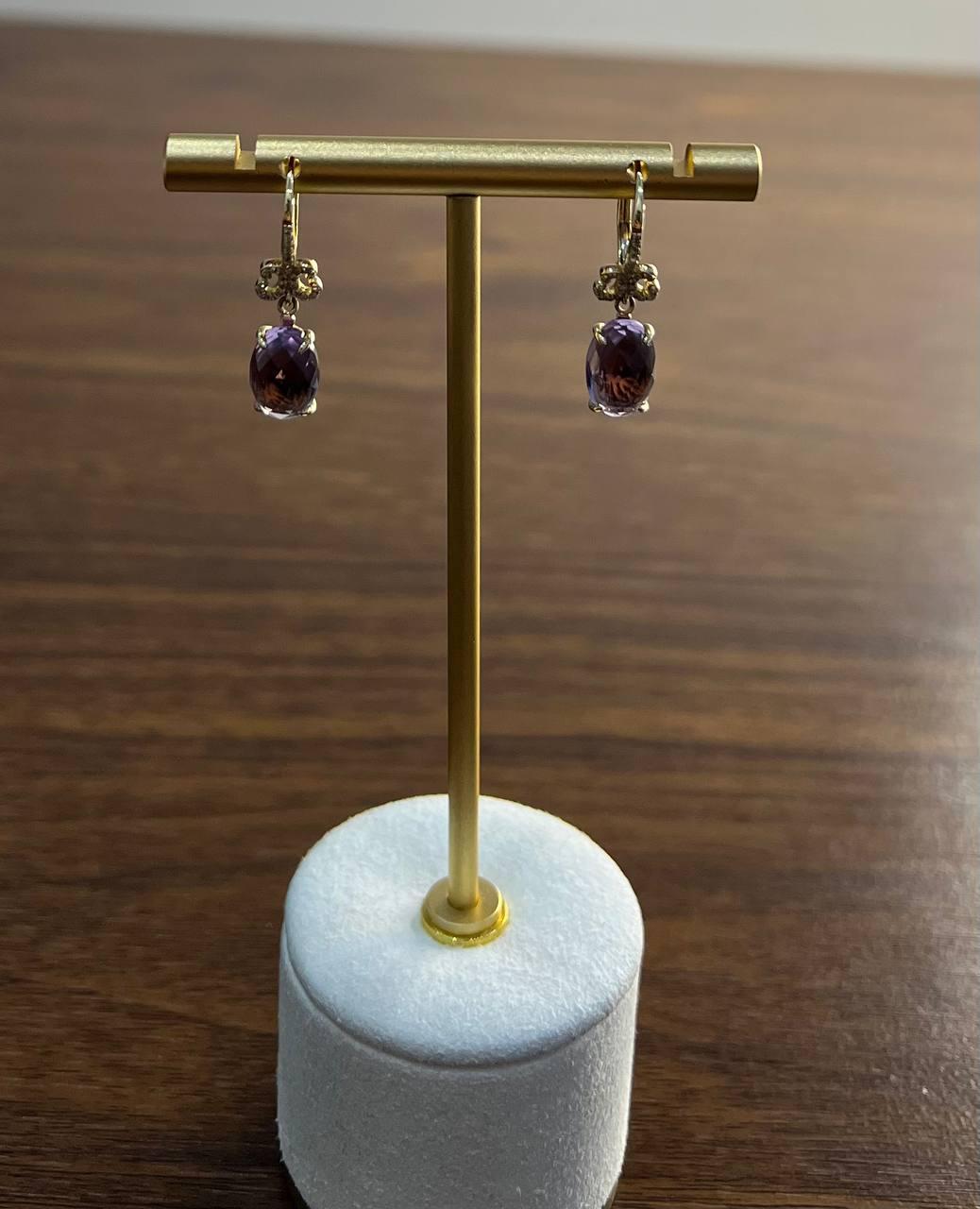 A stunning pair of earrings crafted in 14k gold.
Each earring features a captivating purple amethyst, elegantly suspended from a delicate gold setting. The stones are faceted, allowing them to catch and reflect light beautifully. Above the