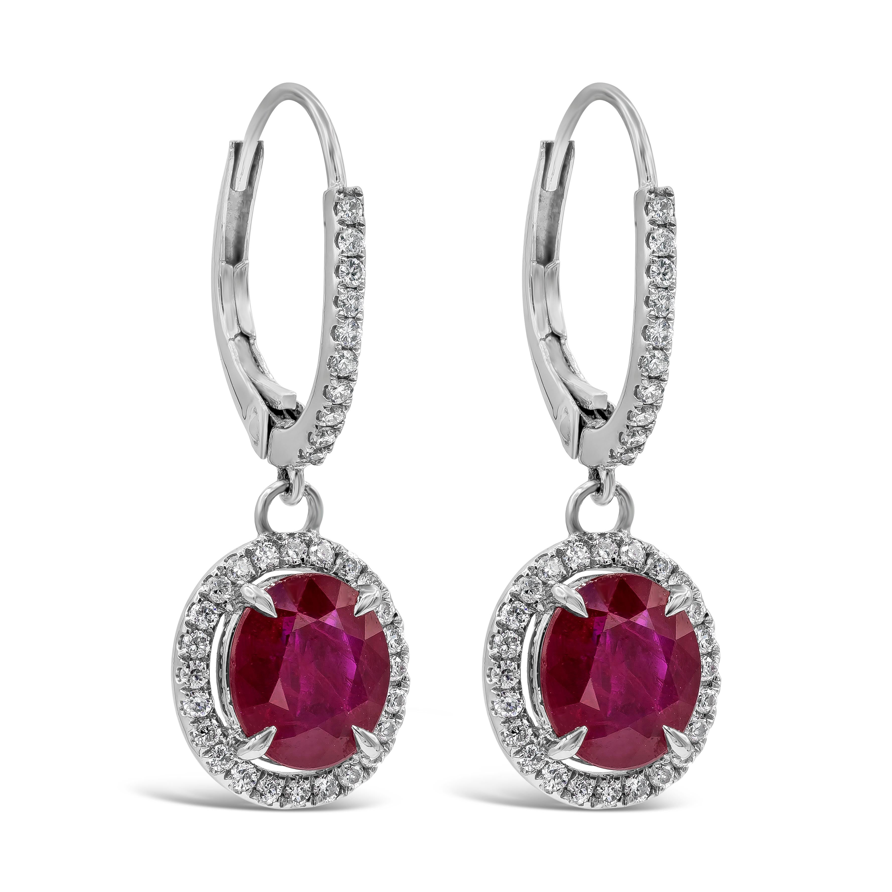 A chic and charming pair of dangling earrings showcasing two oval cut red rubies weighing 2.83 carats total, Surrounded by a row of brilliant round diamonds in a halo design weighing 0.29 carats total. Suspended on a diamond encrusted lever back,