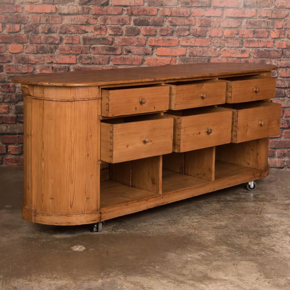 The warm natural pine and curved ends of this freestanding island will create an inviting space to gather in one's kitchen. This unusual oval pine grocer's cabinet with six drawers on one side and paneling on the ends and front originally served as