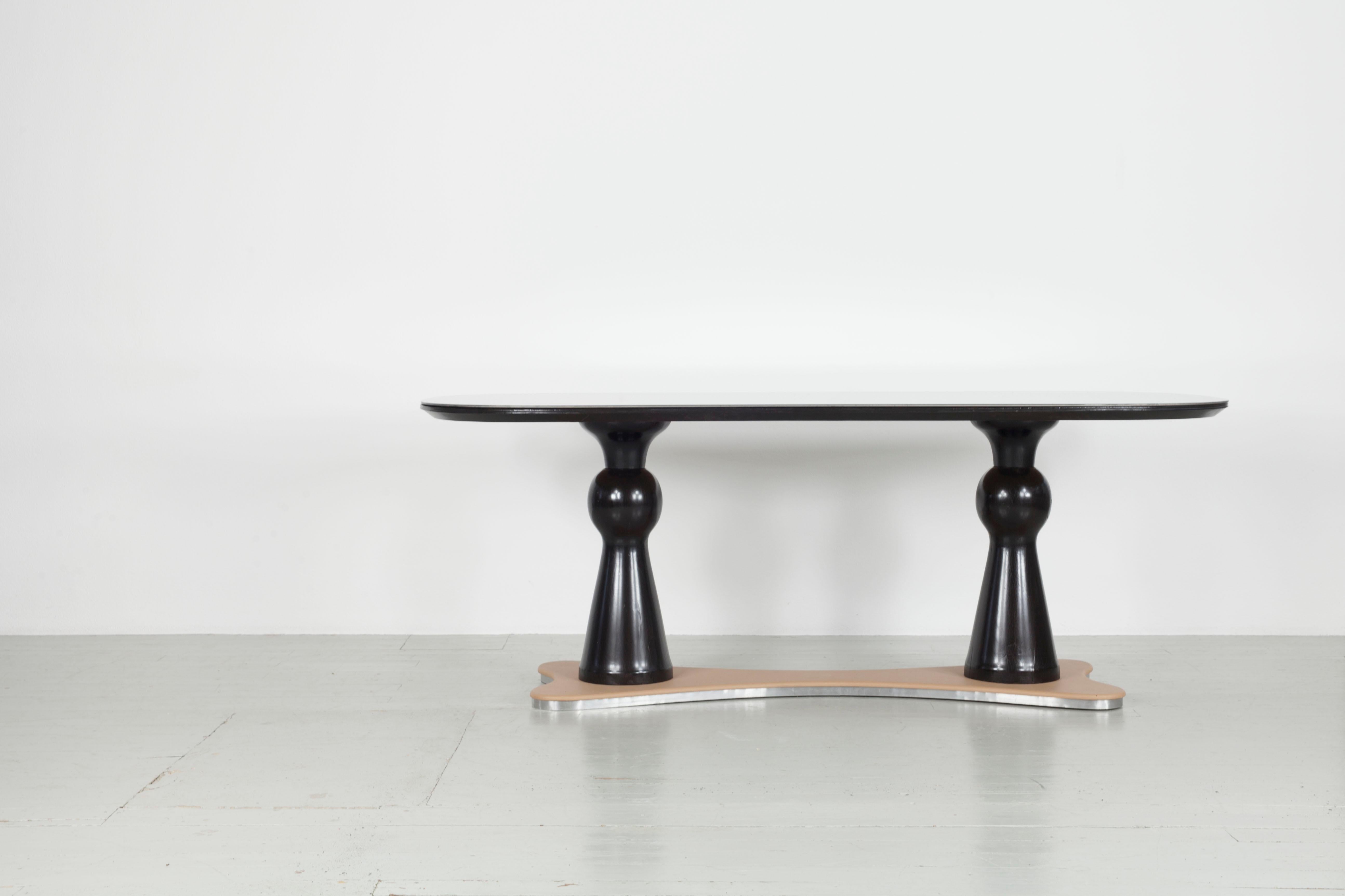 This dining table was made in Italy in the 1940s. The oval table top with grey glass overlay sits on two unusual, dark-stained wooden table legs. These table legs are placed on a table base covered with imitation leather. The table base is framed by