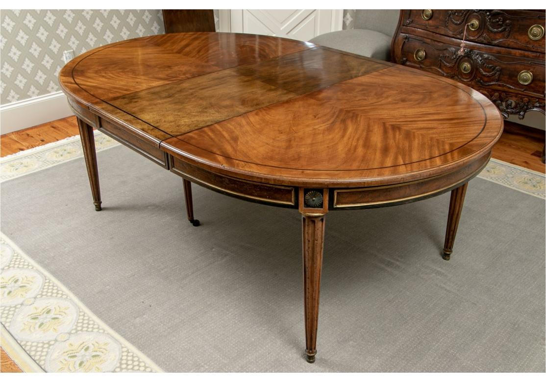 A handsome Directoire Style Dining Table. Oval dining table with beautiful book matched top, carved edge and black string inlaid banding. The ebonized apron with gilt molded panels, and brass rosettes above the fluted tapering legs, all in an