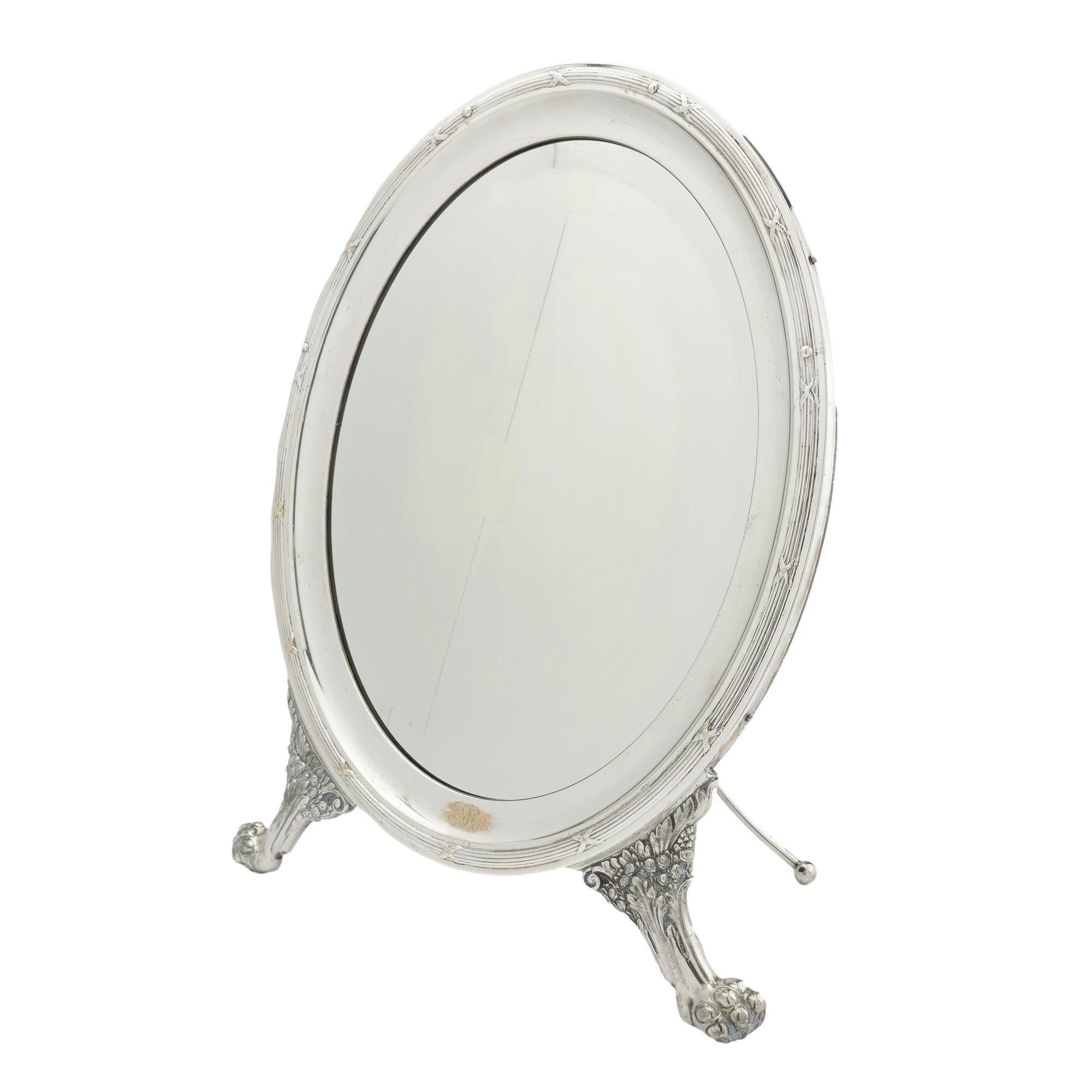 A generously sized oval shaped dressing mirror in silvered brass with an easel back and lion's paw feet. The mirrored glass has a beveled edge and the back is a single board mahogany panel fitted with a hinged silver plated easel stand. The bottom