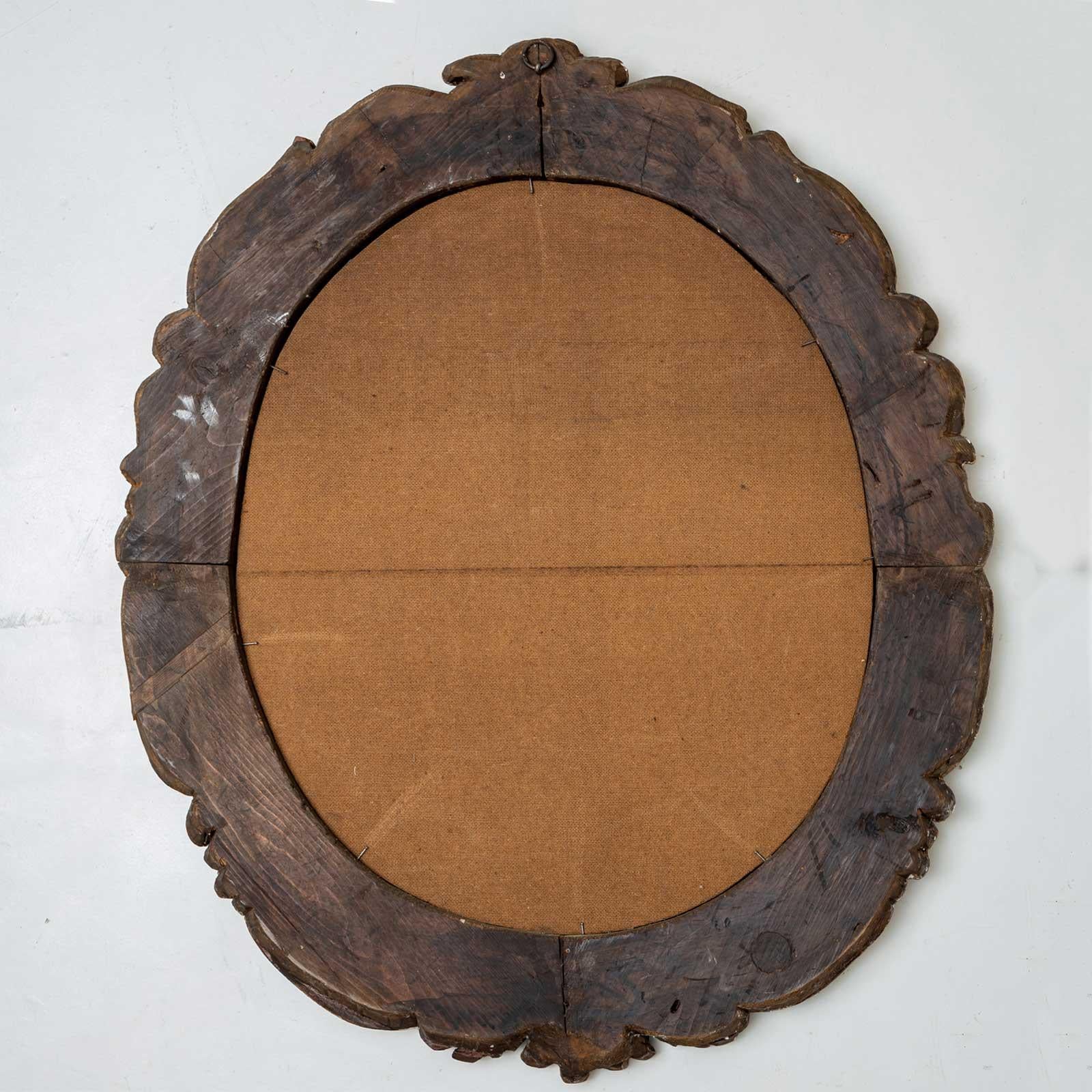 Grand miroir mural ovale dans un cadre en bois sculpté et patiné d'or avec des volutes et des décorations de feuilles. La dorure est frottée par endroits et le Bolus transparaît.