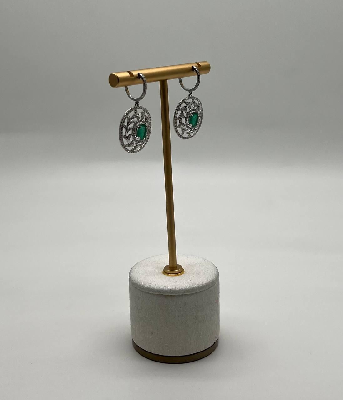 Un sofisticato paio di orecchini con intricati disegni circolari in oro bianco 14 carati. Ogni orecchino è ornato da uno smeraldo verde centrale, che aggiunge un tocco vibrante. Gli orecchini sono sospesi da una delicata cerniera che offre una