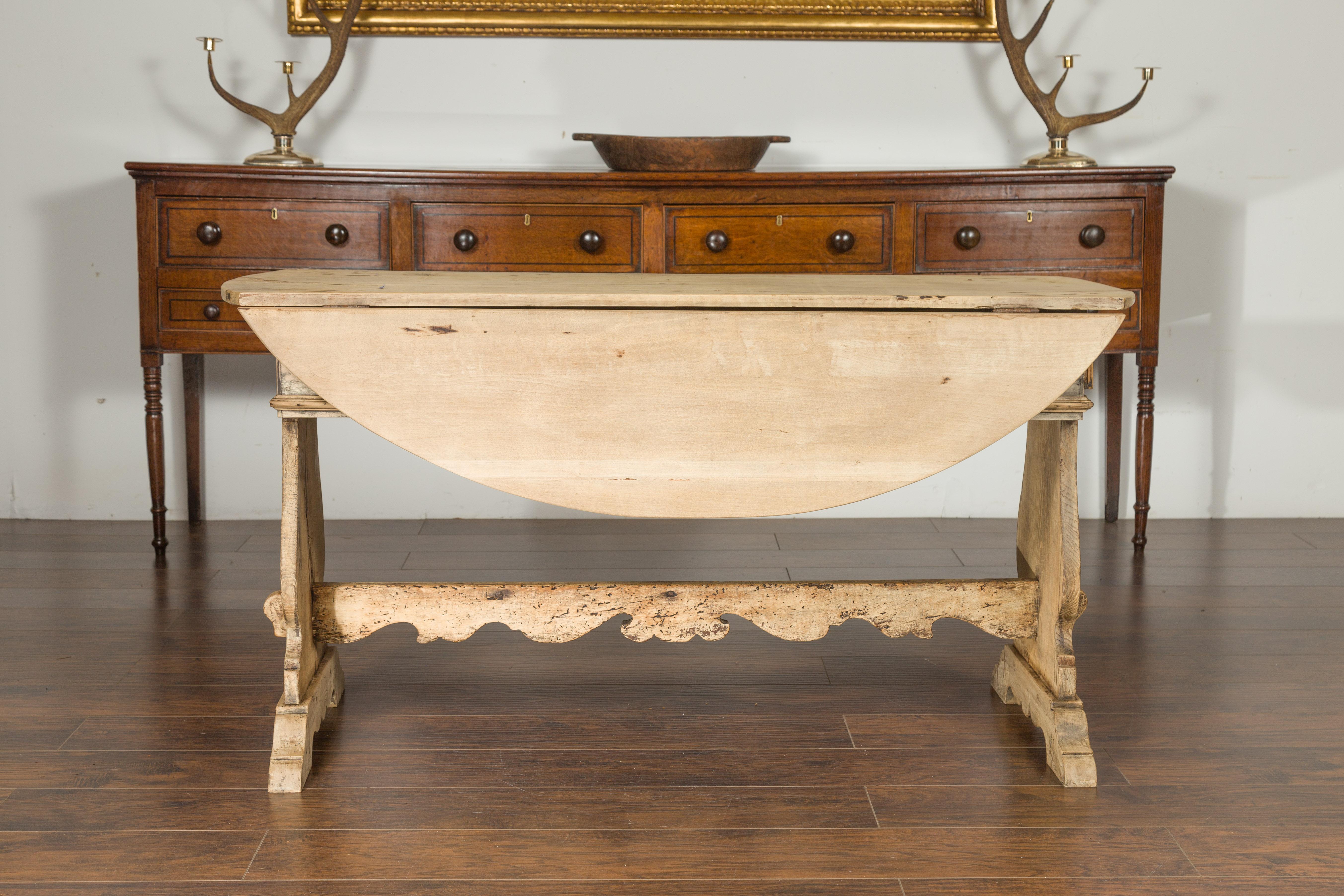 An Oval Italian bleached walnut drop-leaf table from the early 19th century, with trestle base and lateral drawers. Born in Italy during the early years of the 19th century, this Italian table features an oval drop-leaf top. The top sits on a