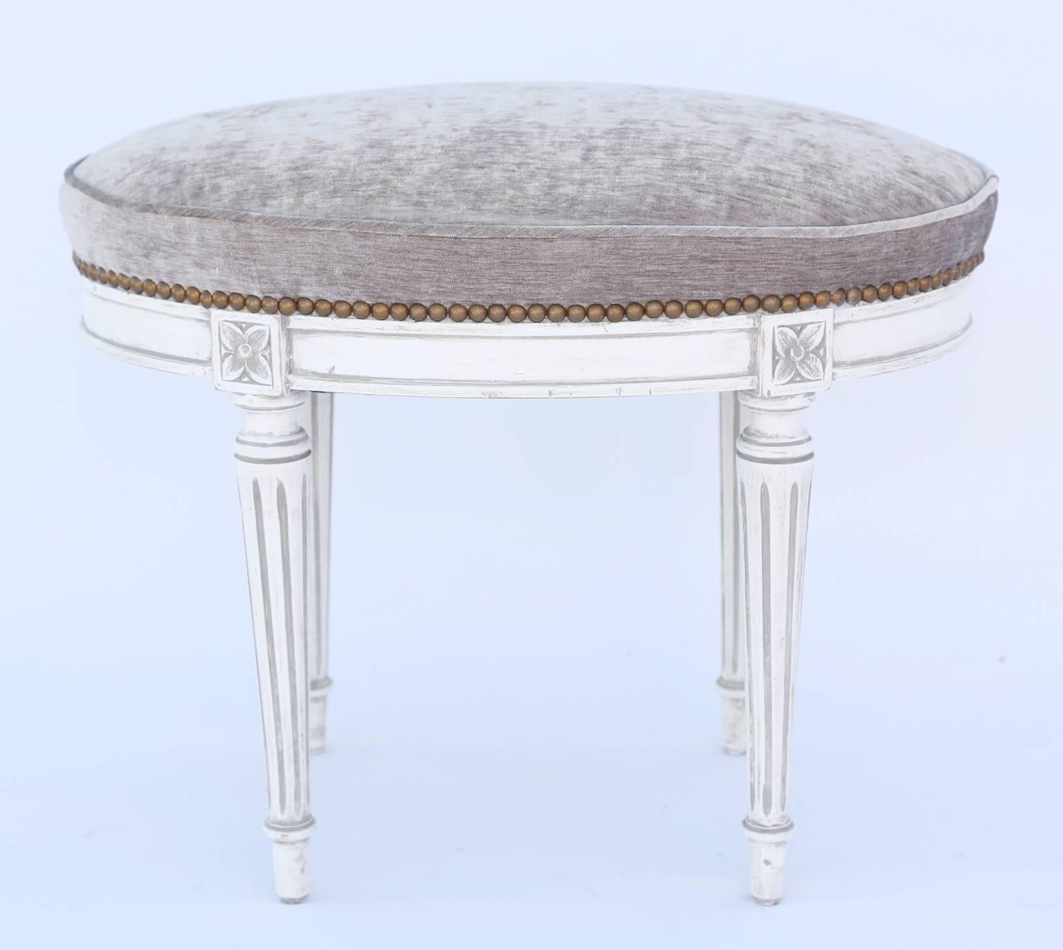 Oval stool, having a boxed seat in mushroom chenille with nailheads, on white painted frame showing natural wear, on fielded apron, raised on round, tapering, fluted legs, ending in touipe feet.

Stock ID: D1338.