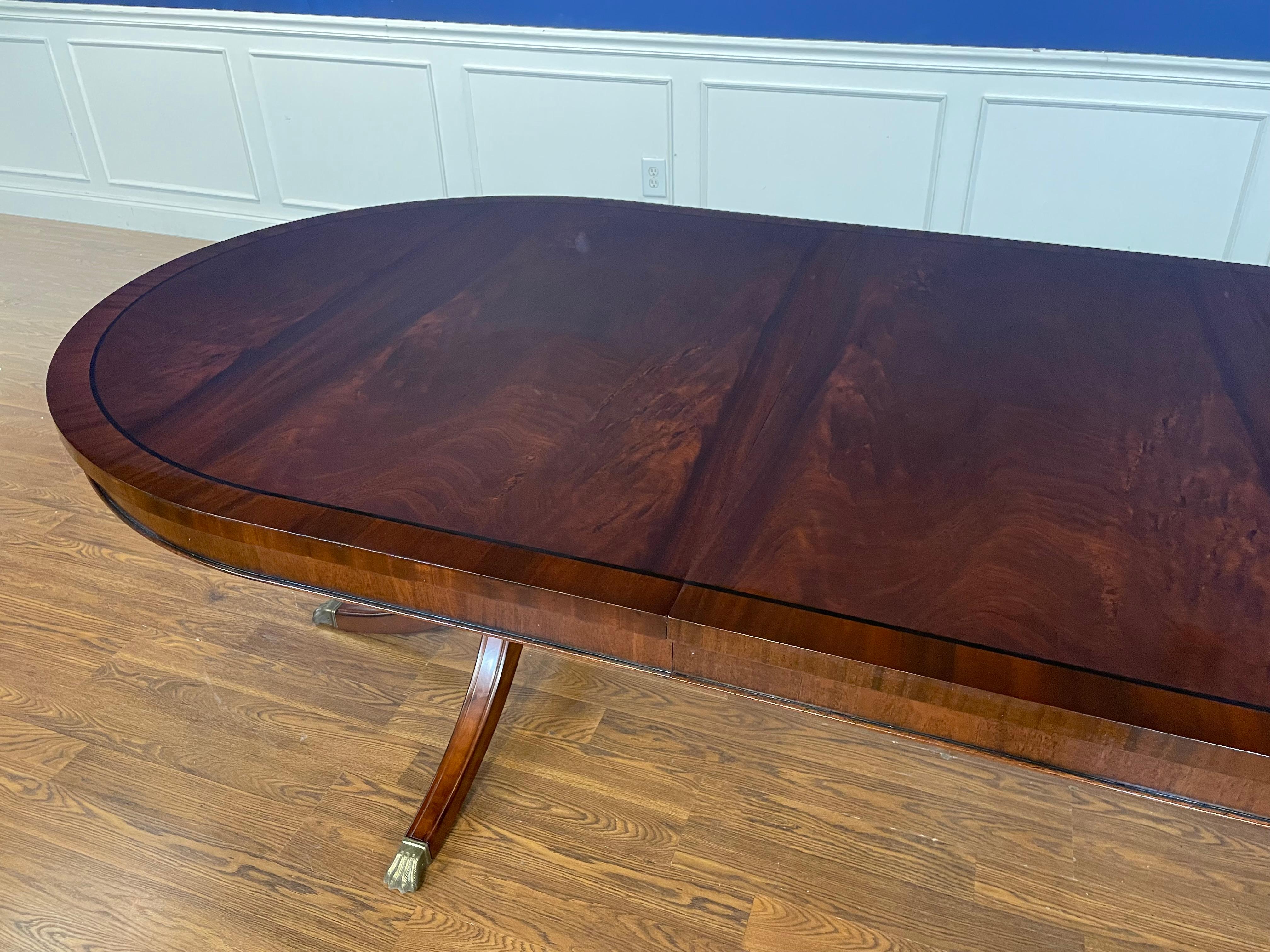 This table features an oval shape with a swirly crotch mahogany field with a straight grain mahogany border. It has two classic Sheraton style pedestals with fluted and tapered legs which terminate with antique brass lions paw feet. It has a hand