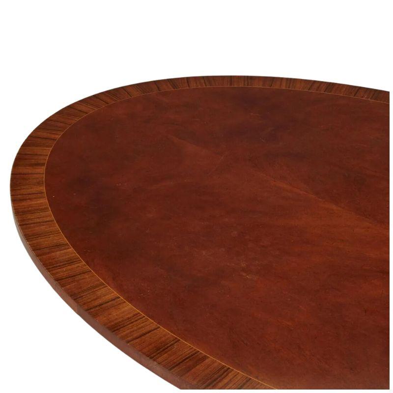 An oval mahogany pedestal breakfast table with a banded edge of a contrasting wood grain.  The classic dining table is a beautifully shaped oval with a turned center pedestal supported by four splayed legs ending in brass paw feet on casters.  From