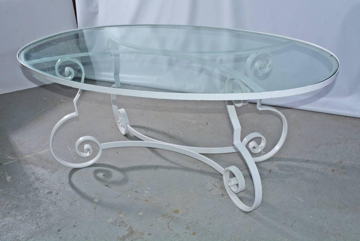 The oval midcentury patio or porch dining table has an iron base and glass top. The base is designed with baroque scroll legs and curved braces that match the curved braces supporting the top. The table is painted white.