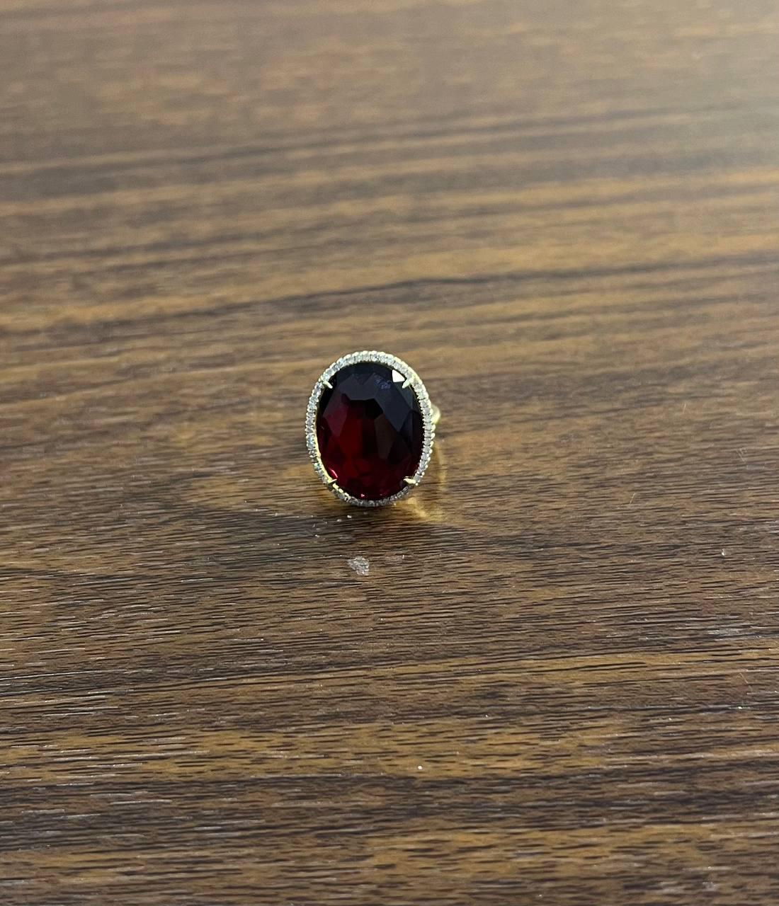 A striking ring featuring a large oval-shaped red garnet. Crafted with 14K Yellow Gold, the ring showcases a vibrant red pink cabochon stone at its center. The stone is encircled by a halo of small, sparkling diamonds, enhancing its vivid color and