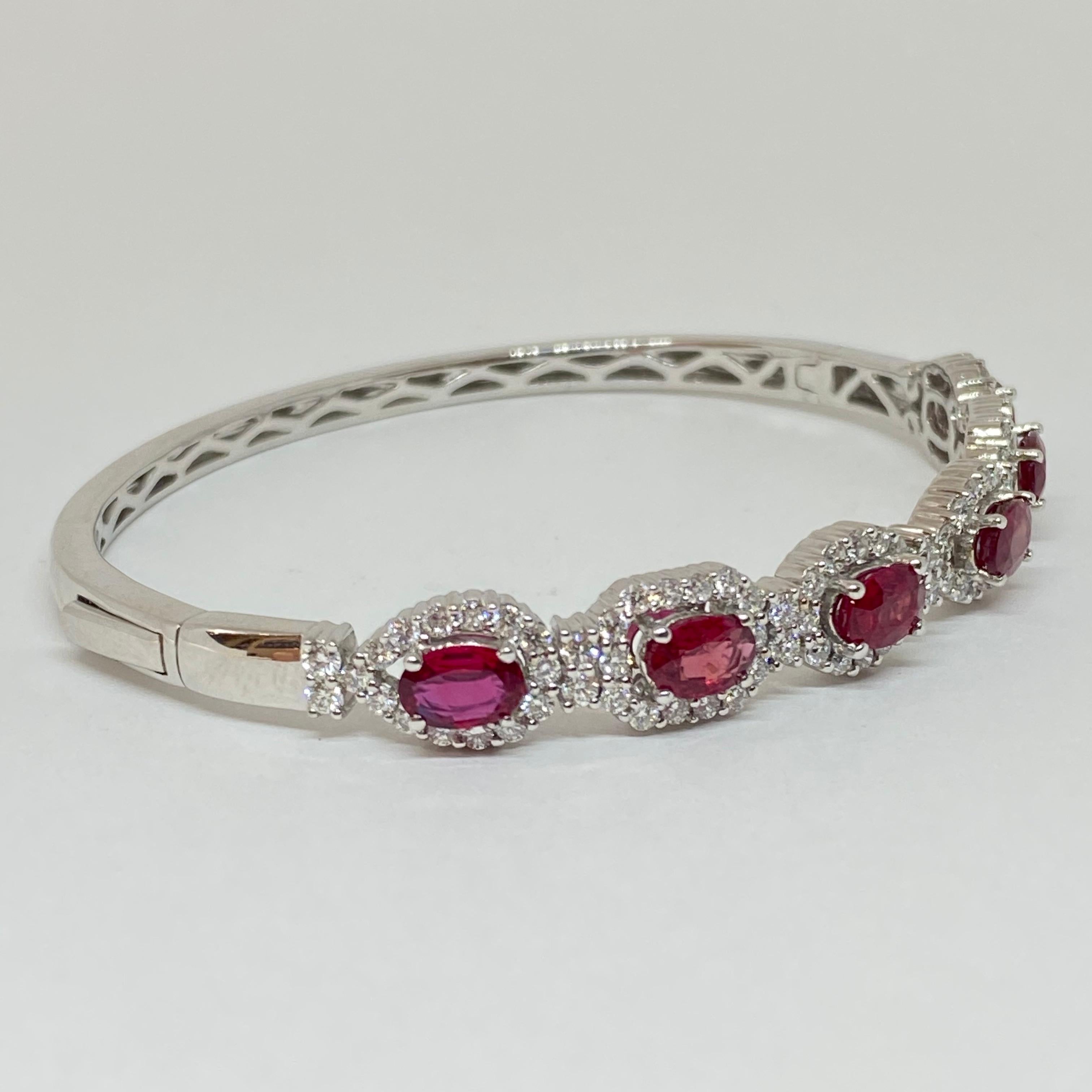 Brand new lovely oval ruby bangle bracelet surrounded by diamond halos, designed in 14 karat white gold. Six vibrant natural oval rubies are set on side with accenting diamonds. Polished white gold bangle with secure button clasp. The rubies have a