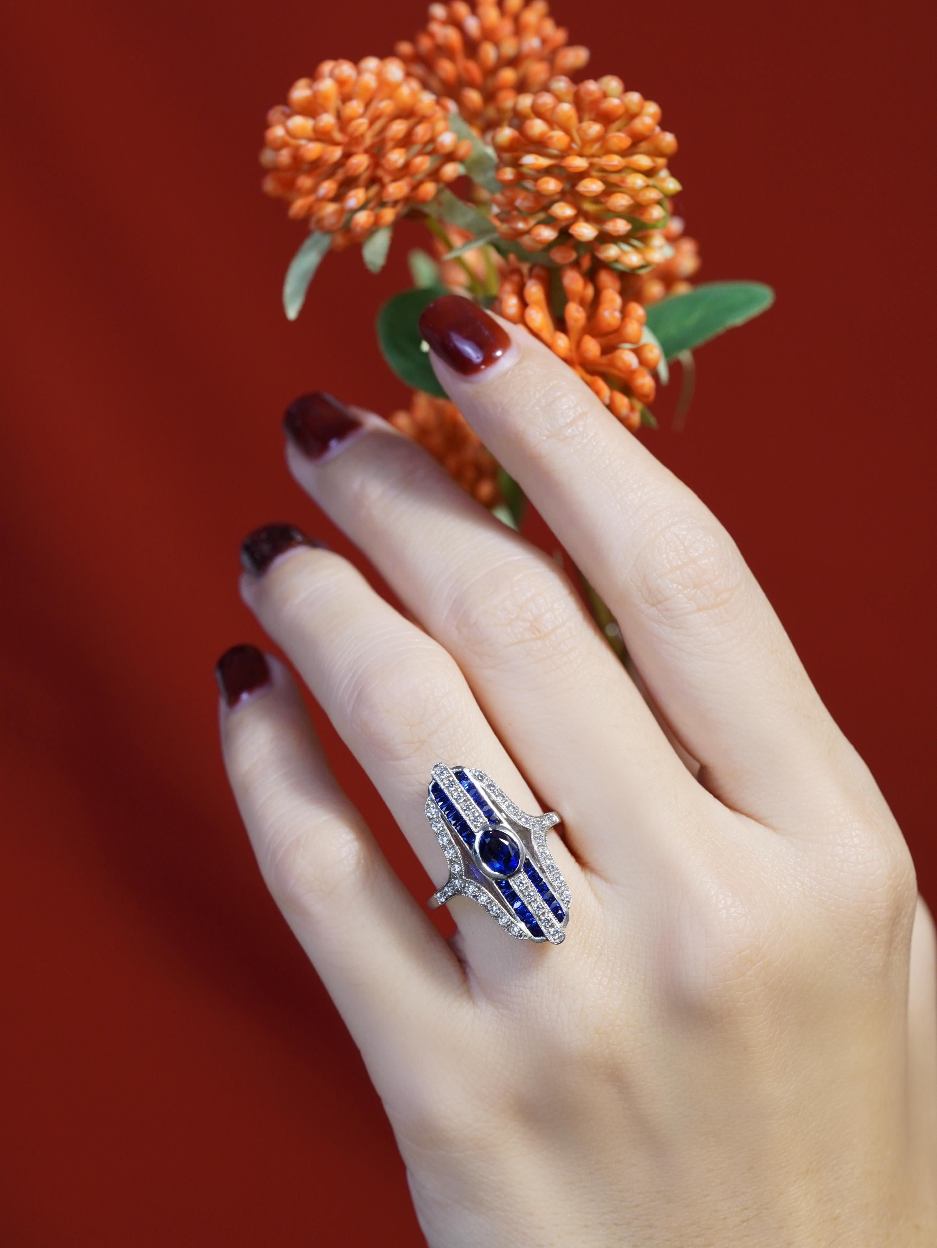 Dieser atemberaubende Art-Déco-Cocktailring zeigt einen ovalen Saphir in Lünettenfassung in der Mitte, flankiert von Saphiren auf beiden Seiten, die für ein reiches, lebendiges Aussehen sorgen. Die Schultern sind elegant mit funkelnden Diamanten