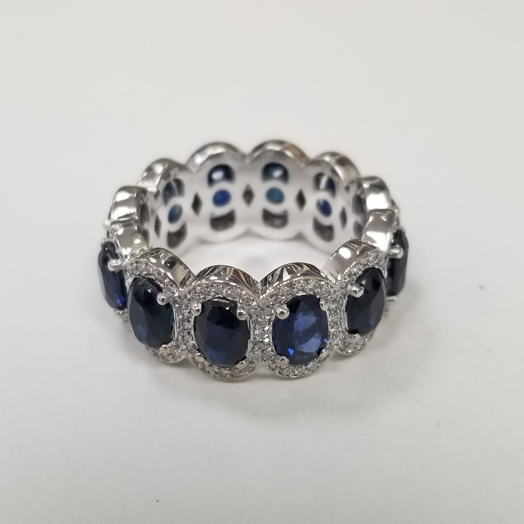 A breathtakingly beautiful diamond wedding band featuring 12 oval cut blue sapphires set in 14k white gold surrounded by a diamond halo. This seamless eternity band is expertly crafted surely will catch eyes. It measures 9.5mm wide. This classic